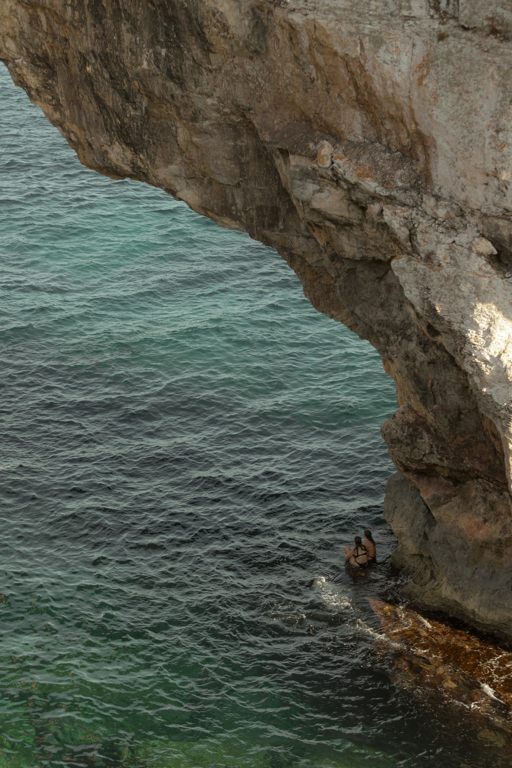 Rock cliff over turquoise ocean with two people sitting on the rocks near the water's edge.