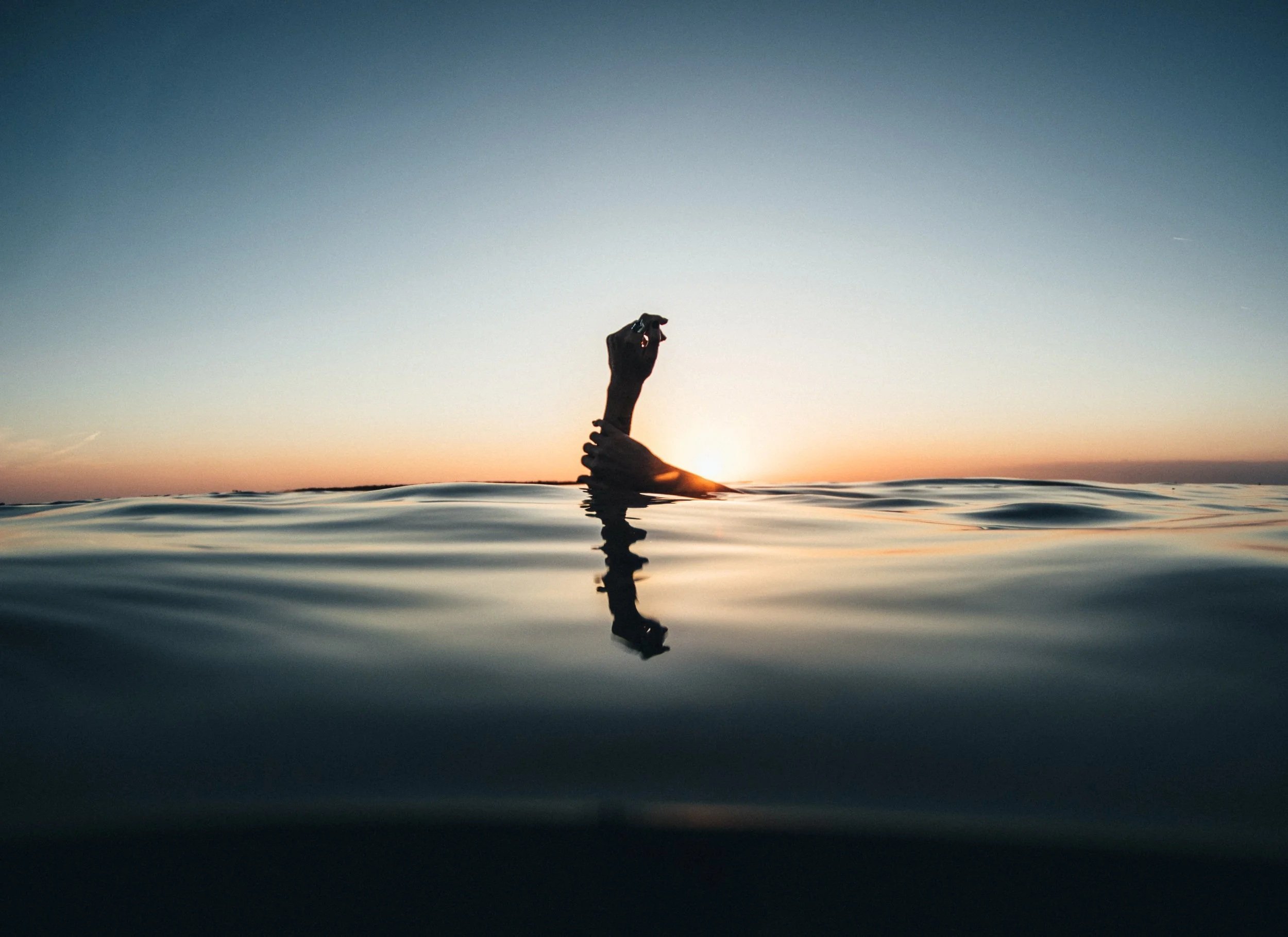 A person’s hand out of the water during sunset or sunrise, with a calm ocean and sky in the background.