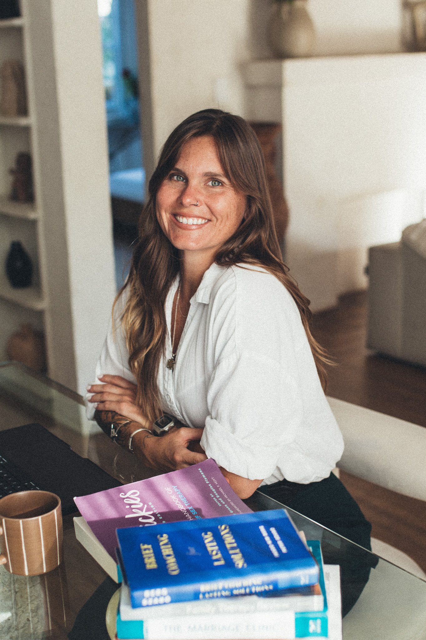 A young woman with long brown hair and light skin, smiling and sitting at a table. She is wearing a white shirt, and around her wrist are a watch and jewelry. The table has books, papers, and a cup, with a living room visible in the background.