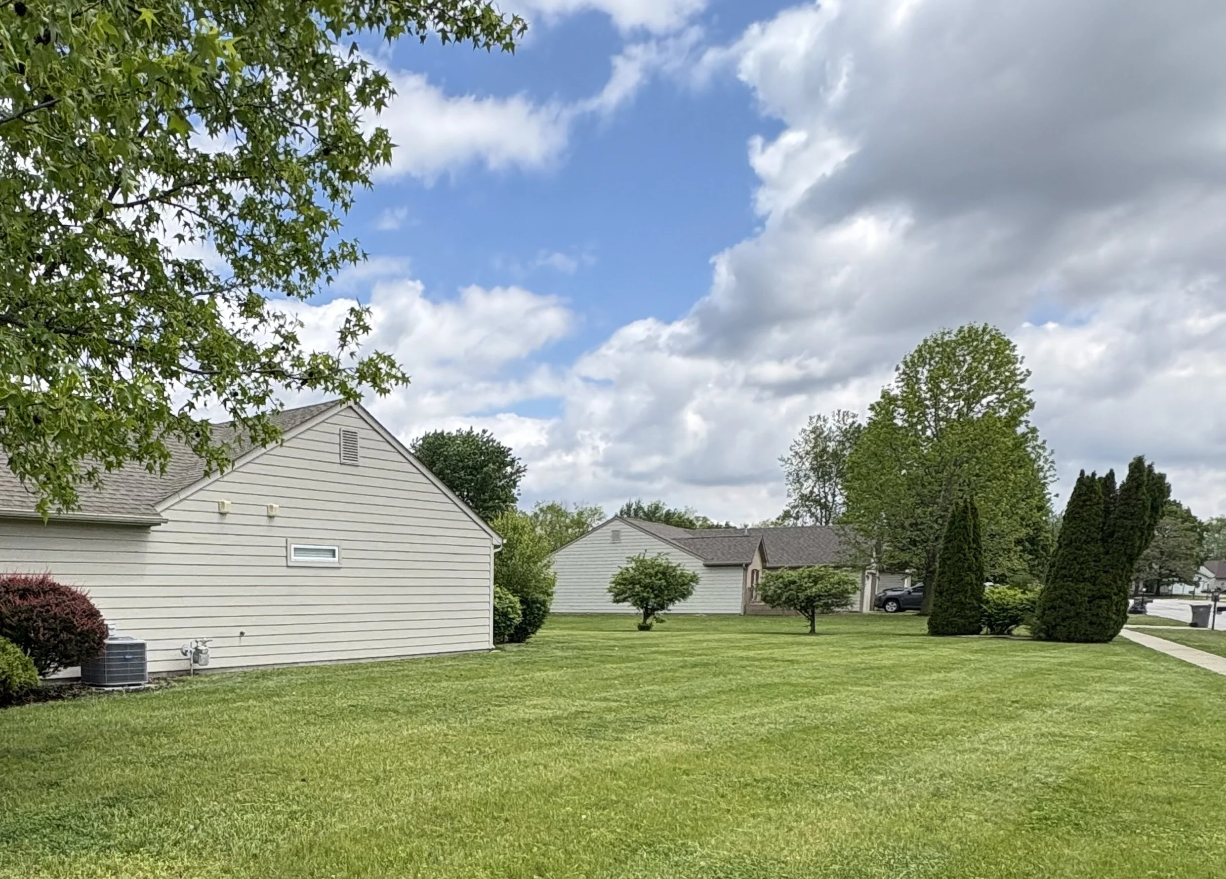 A residential backyard with green grass, multiple trees, and one white house with a gray roof. The sky above is partly cloudy with blue skies visible.