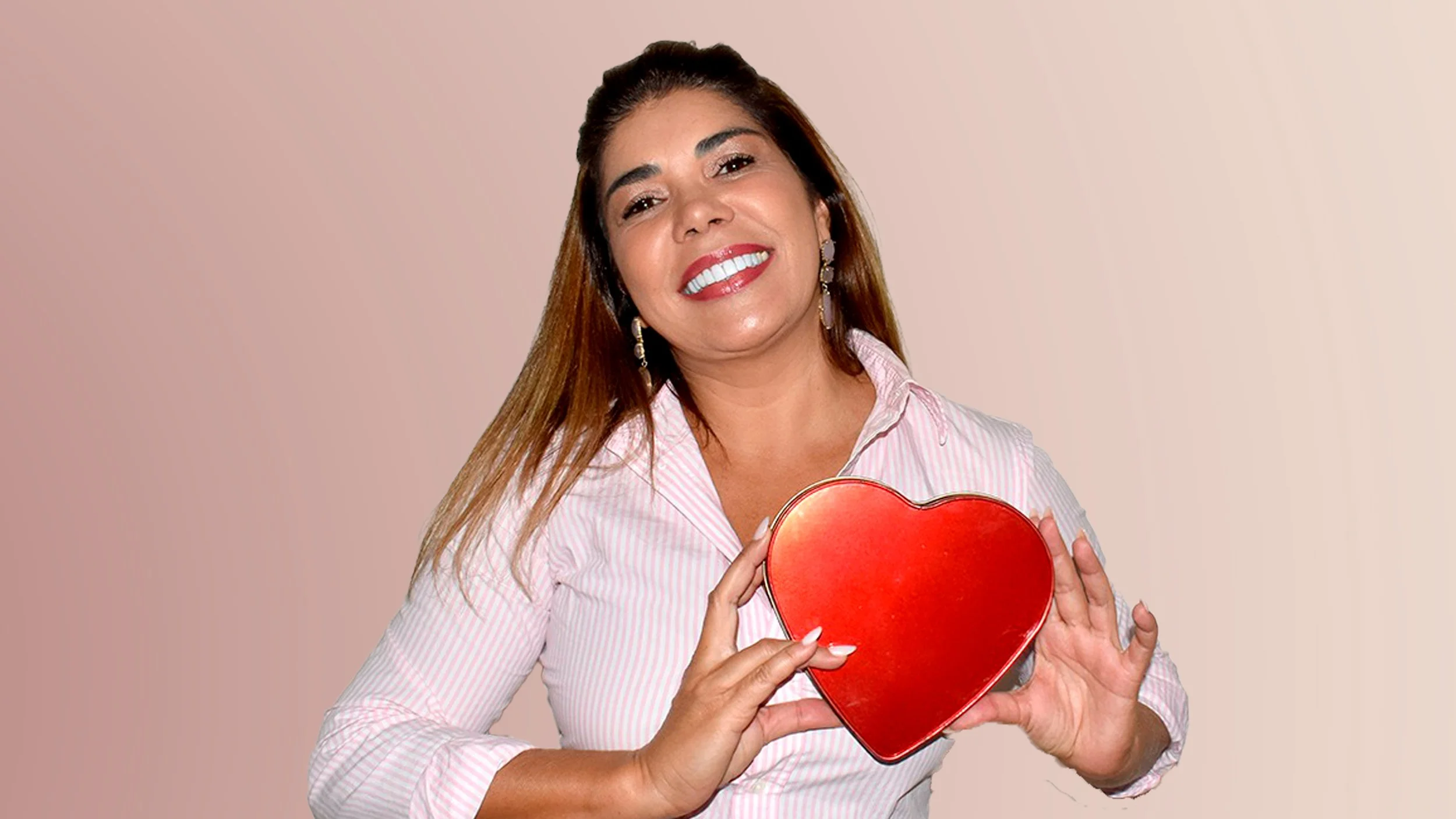 Mulher sorridente com cabelo castanho, usando blusa rosa de listra branca e brincos dourados, cruzando os braços em frente a uma parede branca.
