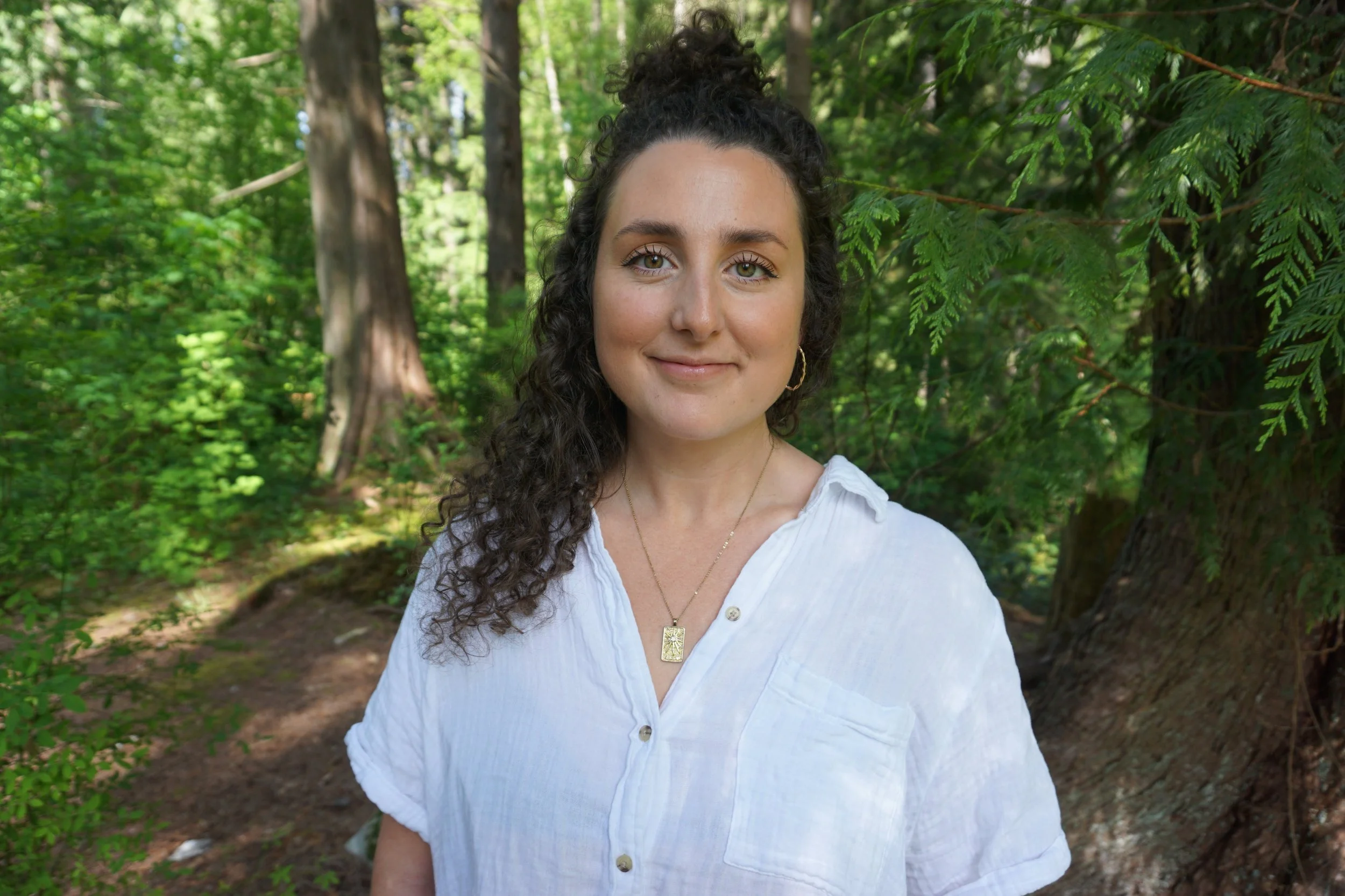 A woman with curly dark hair tied up in a bun, wearing a white top and gold necklace, standing in a lush green forest with trees and foliage in the background.