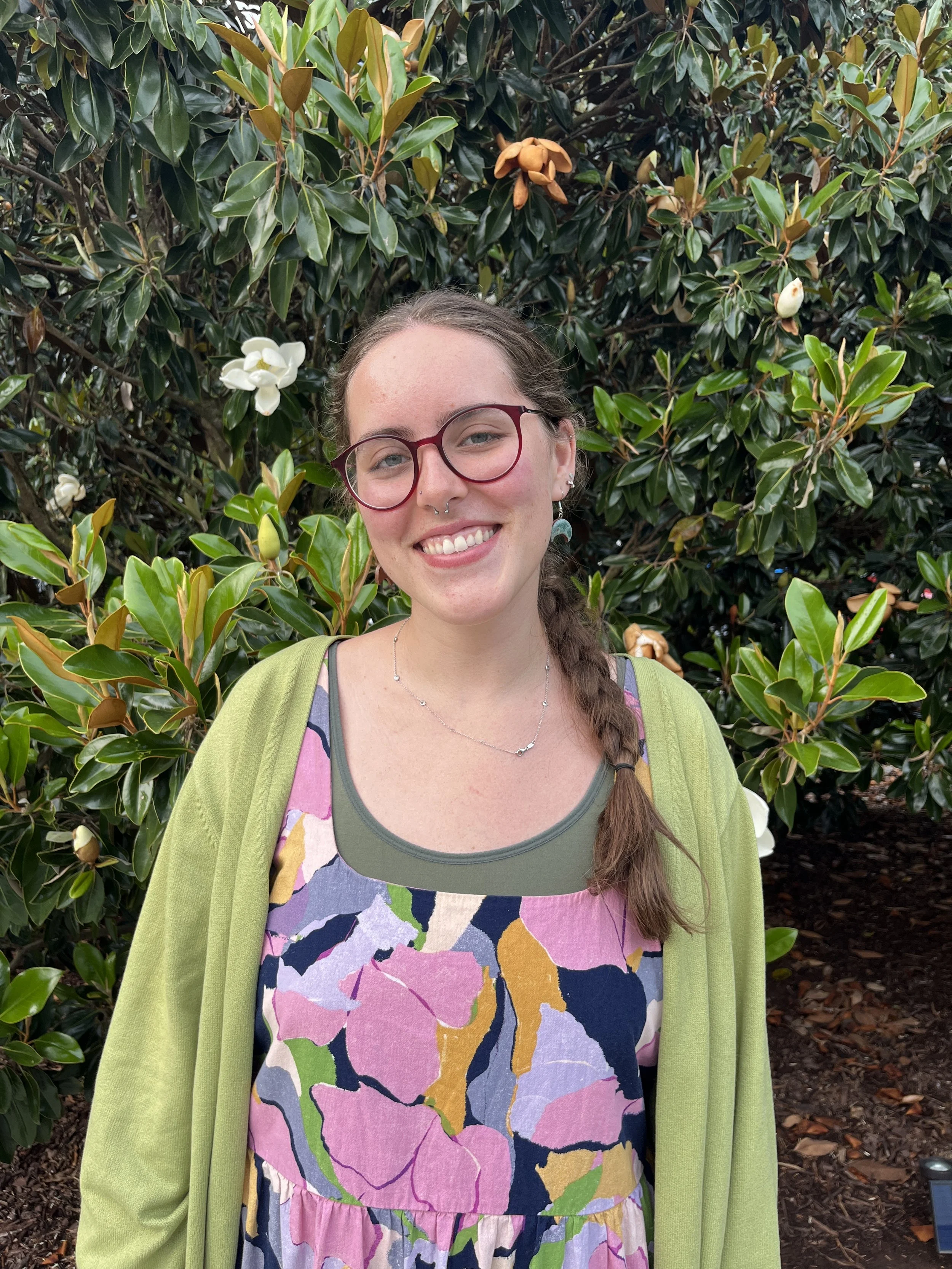 A young woman with glasses and long braided hair smiling outdoors in front of a magnolia tree with white and brown flowers, wearing a colorful dress and green cardigan.