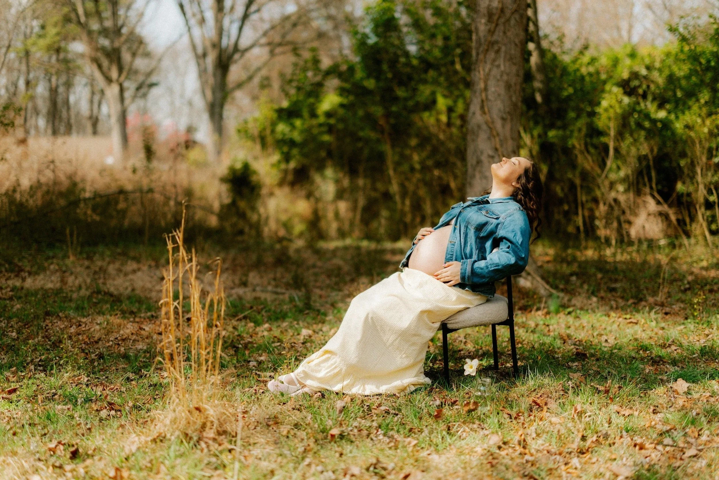 Pregnant woman sitting outdoors in a park, enjoying nature with her eyes closed and a smile, wearing a denim jacket and a long beige skirt.
