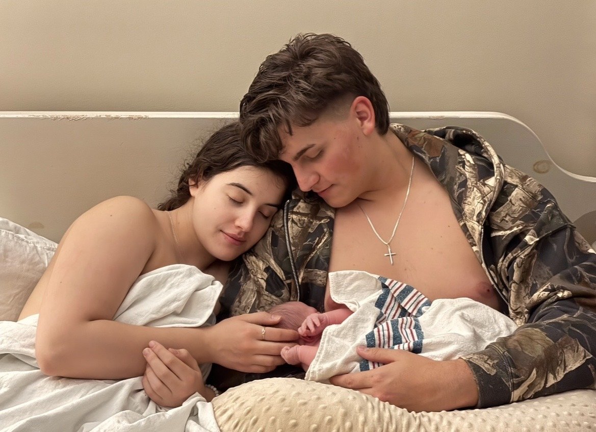 A young woman and a young man are sitting in bed, holding a newborn baby between them. They are resting their heads together, looking at the baby with affection.