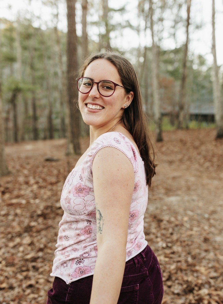 Smiling woman with glasses and tattoos standing in a forest with fallen leaves.