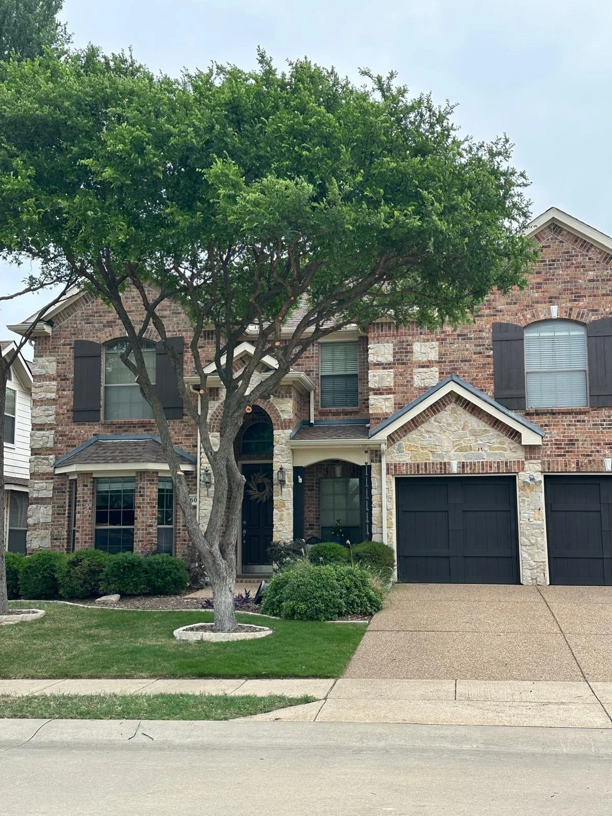 A two-story brick house with black shutters, a small front porch, and a black garage door, with a large leafy tree in the front yard, and bushes and a lawn.