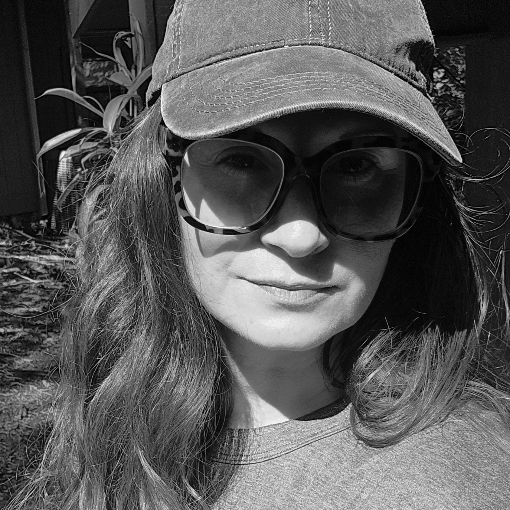 A woman wearing a baseball cap, sunglasses, and a t-shirt with her long hair loose. She is outdoors with foliage in the background.