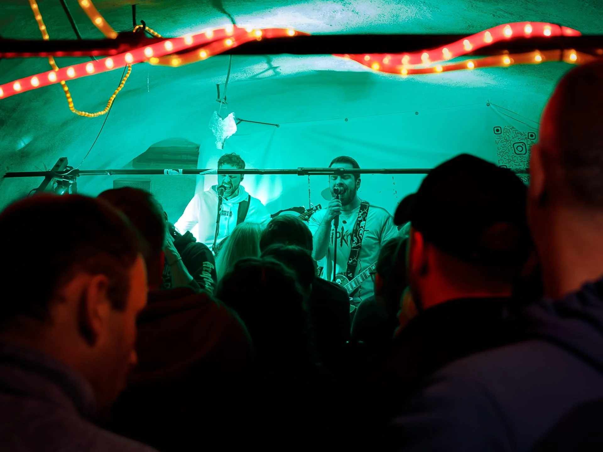 Two men are performing live music on a small stage with colorful lighting, surrounded by a crowd of concertgoers.