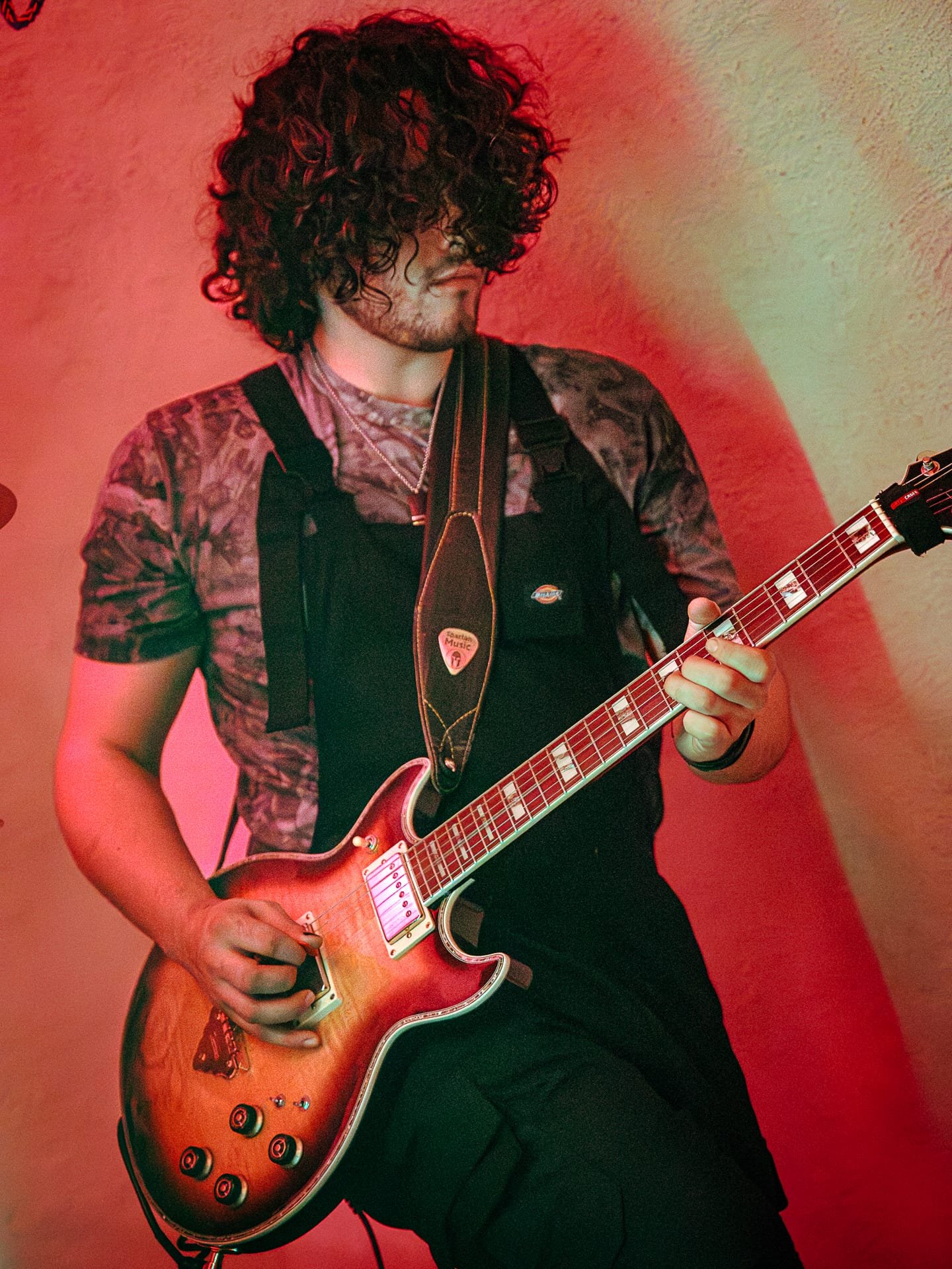 A person with curly dark hair playing an orange sunburst electric guitar against a textured red wall.