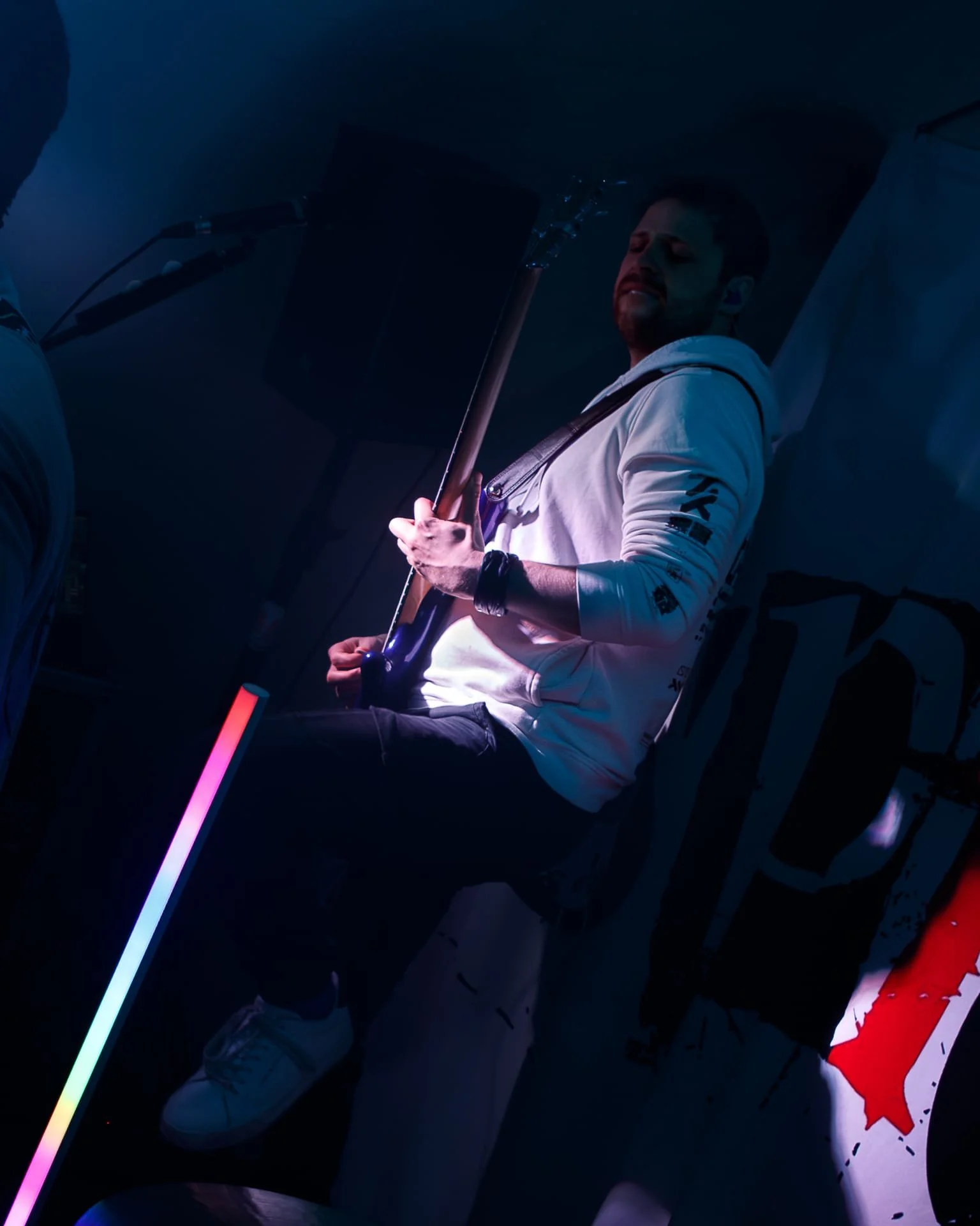 A young man in a white hoodie and jeans playing an electric bass guitar on a dark stage with colorful lighting.