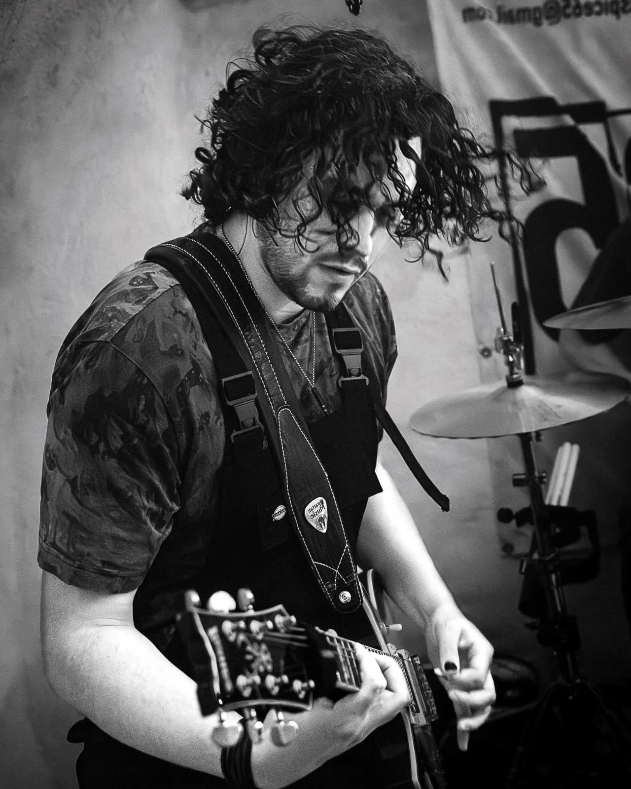 A young man with curly hair playing an electric  guitar while wearing a camouflage shirt and dungarees, surrounded by musical instruments in a black-and-white setting.