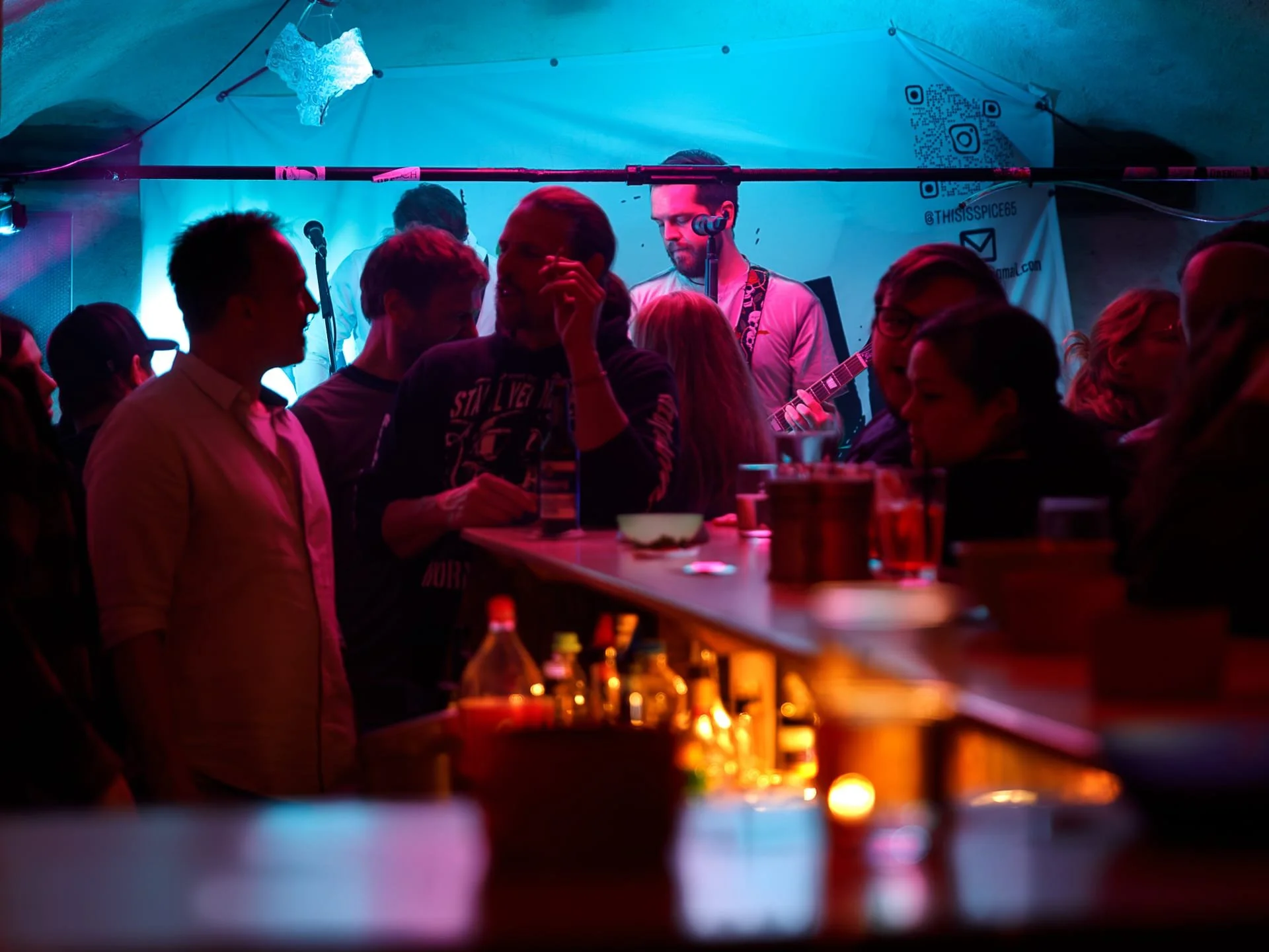 People gathered in a dimly lit bar or nightclub with a live band performing on stage, a guitarist visible, and colorful lighting creating a lively atmosphere.
