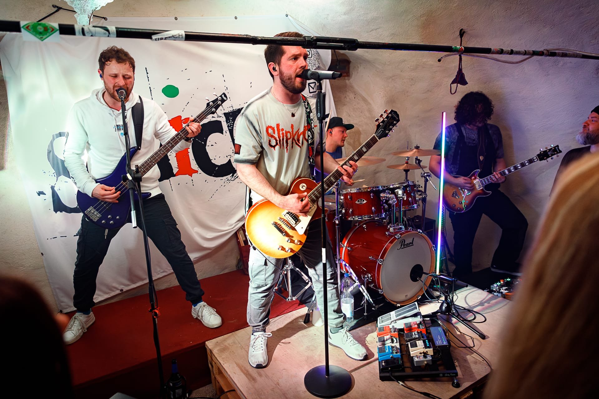 A band performing live on a small stage, with four musicians playing guitars and drums, under colorful lighting, and a white backdrop with a logo or design.