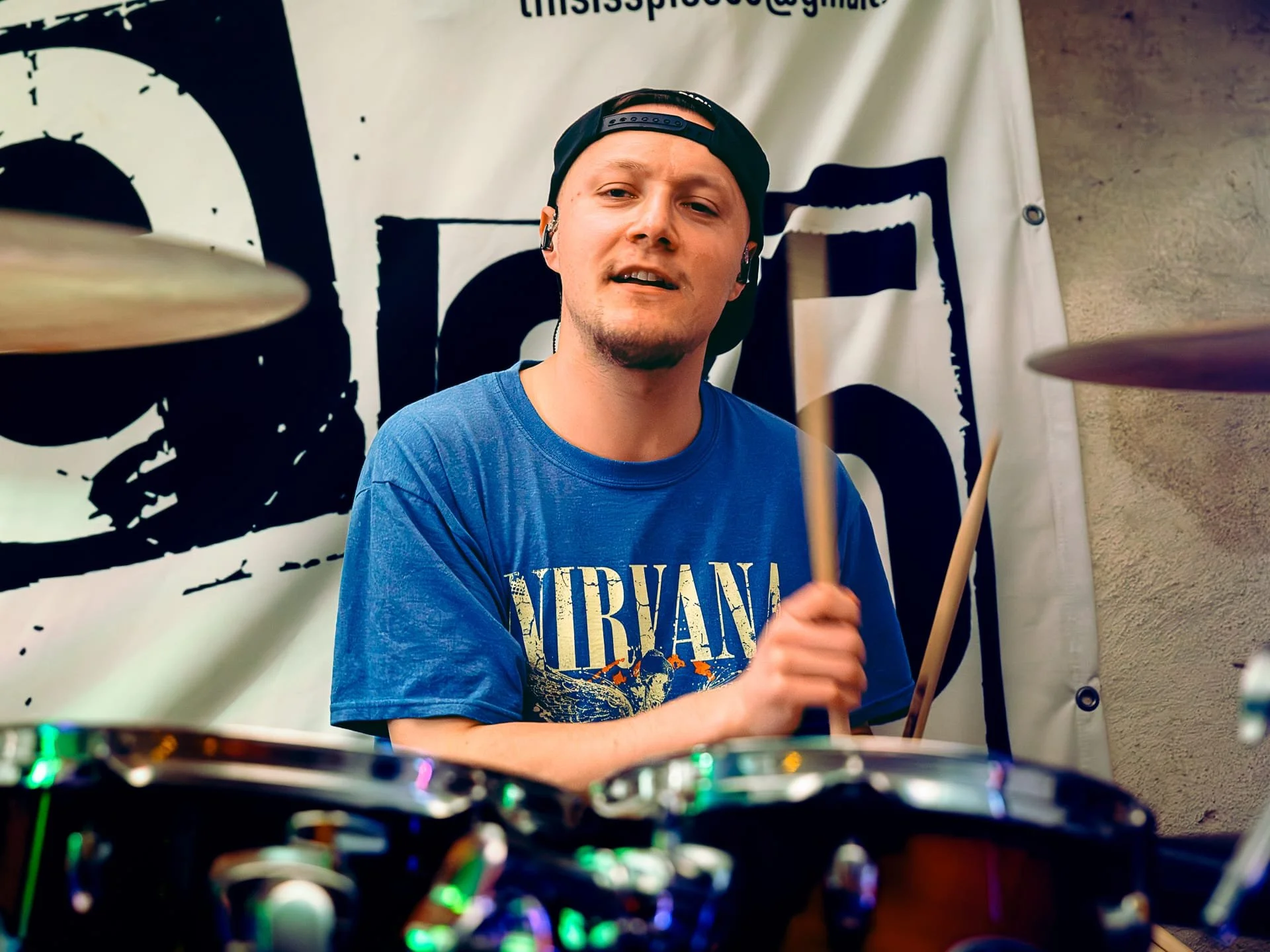A young man wearing a black backward cap and a Nirvana band T-shirt is playing drums during a performance. He is smiling, holding drumsticks, with a Nirvana banner in the background and partial view of drums and cymbals in the foreground.