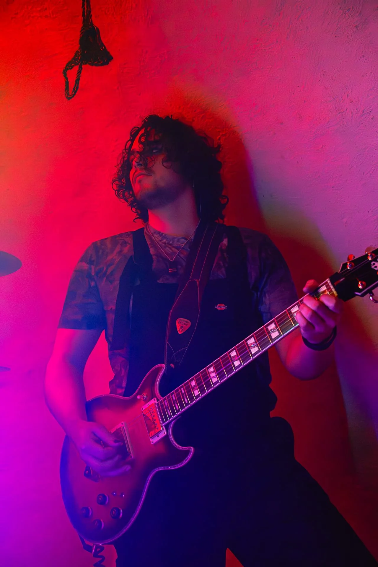 A man with curly hair and a beard playing an electric guitar in a room with pink and purple lighting.