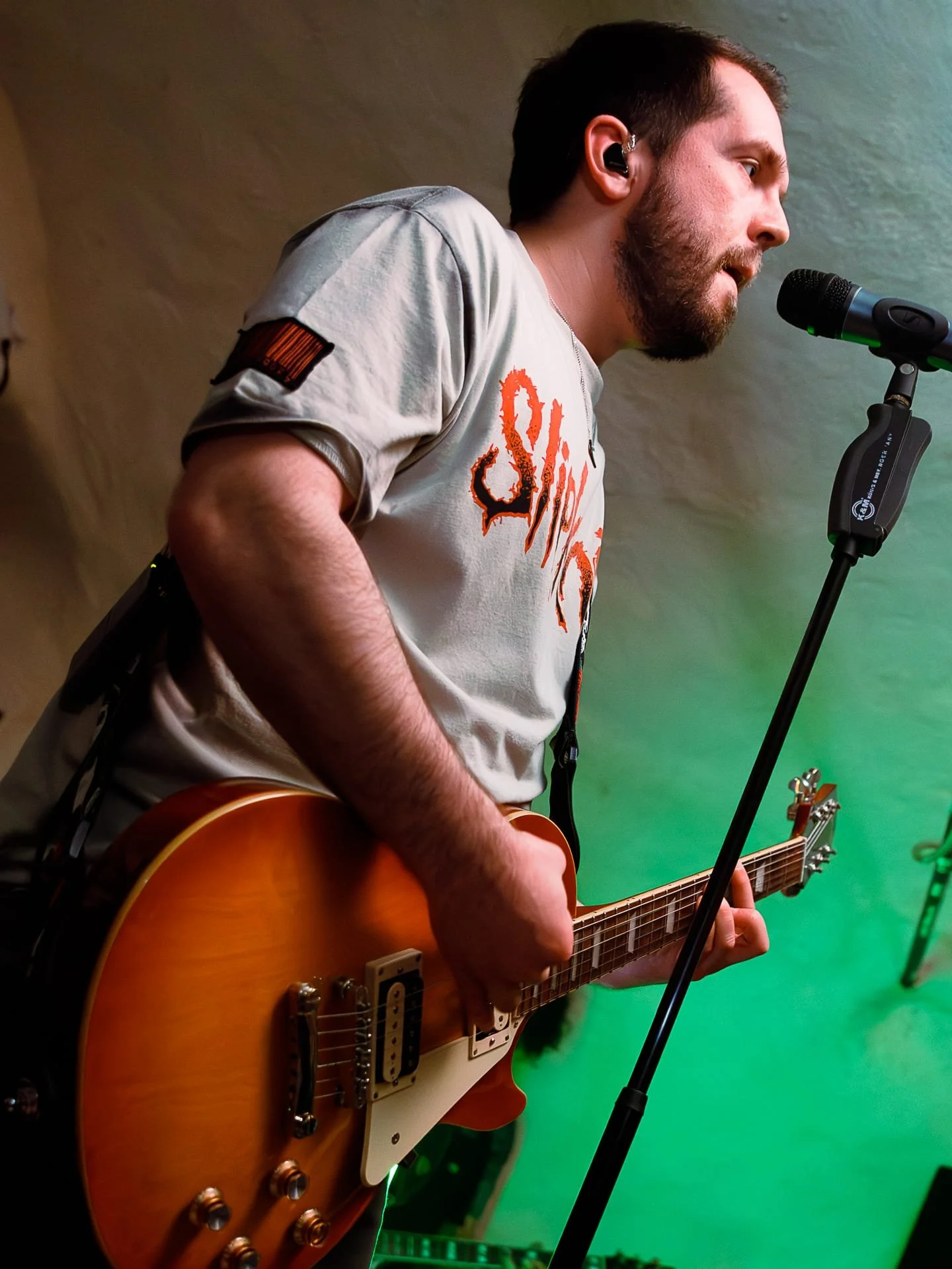 A man with short dark hair, beard, and wearing a white t-shirt, is playing an electric guitar and singing into a microphone, with a green background.