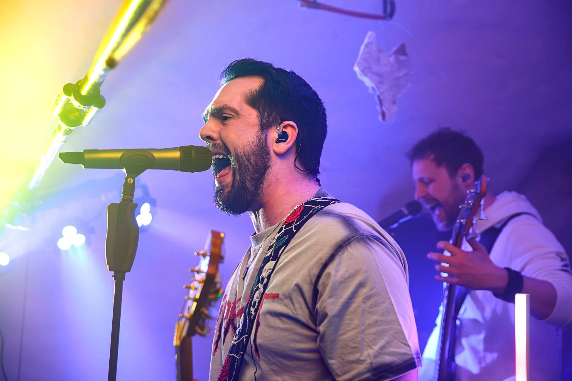 Male singers singing into a microphone on stage, with another musician playing guitar in the background, illuminated by colorful stage lights.