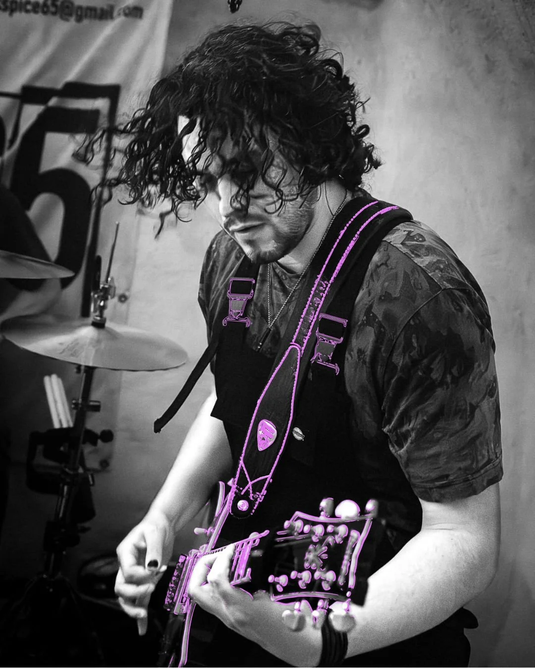 A person with curly hair playing an electric guitar, with parts outlined in purple, in a room with a drum set in the background.
