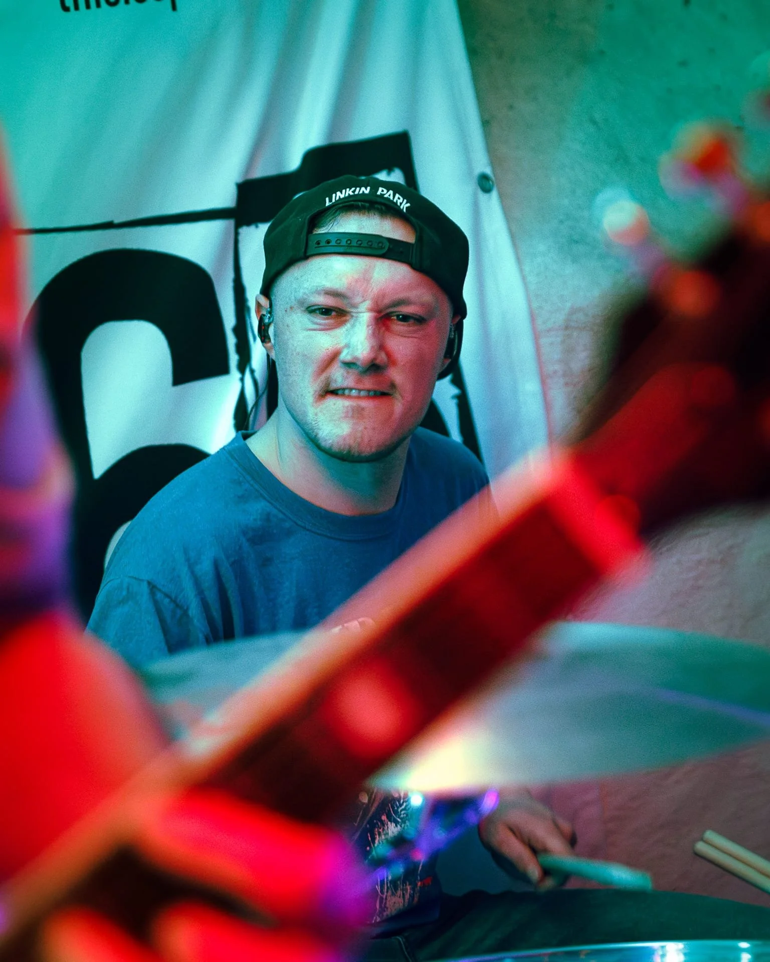 A person with a mischievous or angry expression, wearing a black backward cap reading 'LINKIN PARK,' seated at a drumset , with a blurry colorful foreground element and background, possibly at a social gathering, a concert or party.