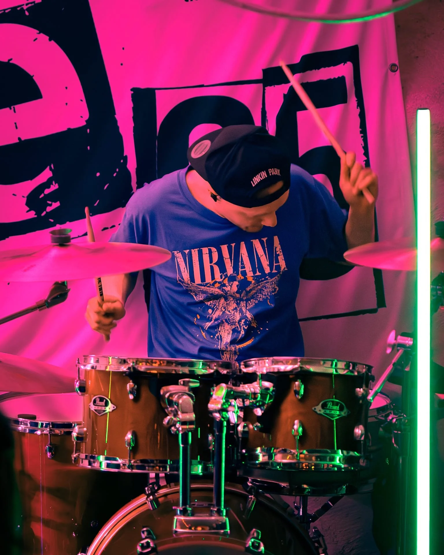 A person wearing a Nirvana t-shirt and a black cap playing a drum set under pink and green neon lights.