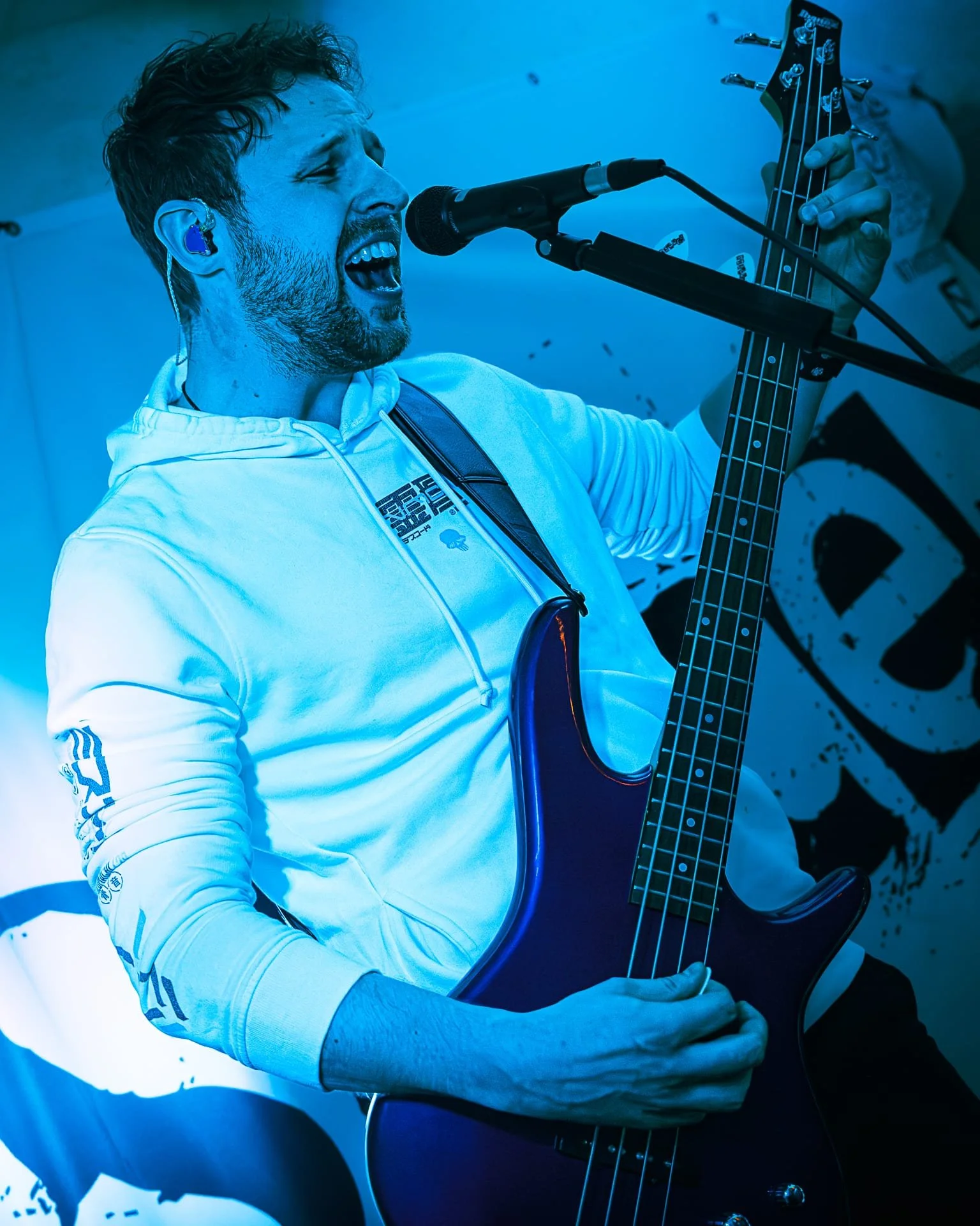 A man passionately singing or shouting into a microphone while playing an electric bass guitar, wearing a white hoodie, with a blue overlay effect.