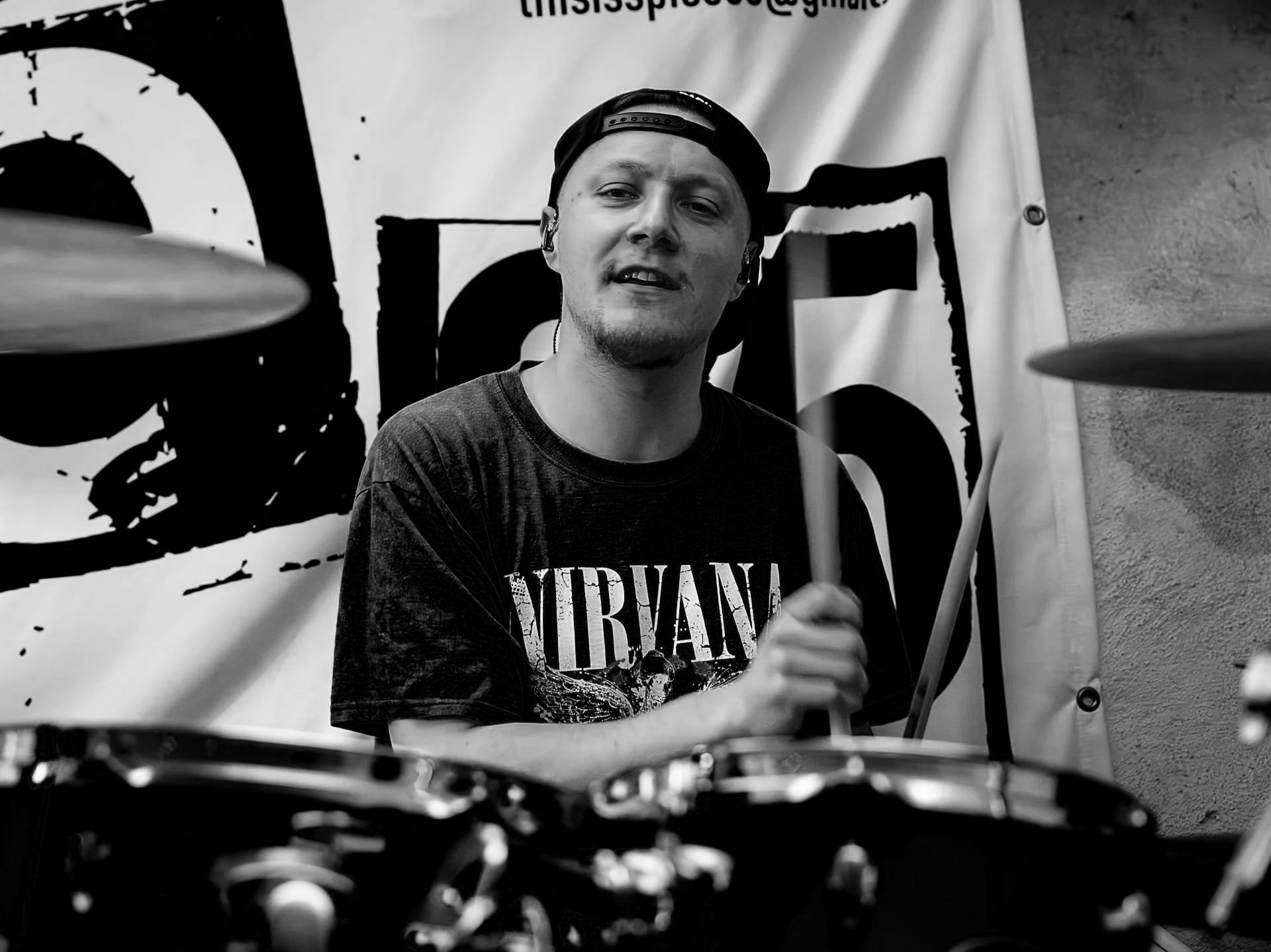 Paddy mit Baseballkappe und Nirvana-Shirt spielt Schlagzeug bei einem Konzert, im Hintergrund eine Fahne mit Spice 65 Druck