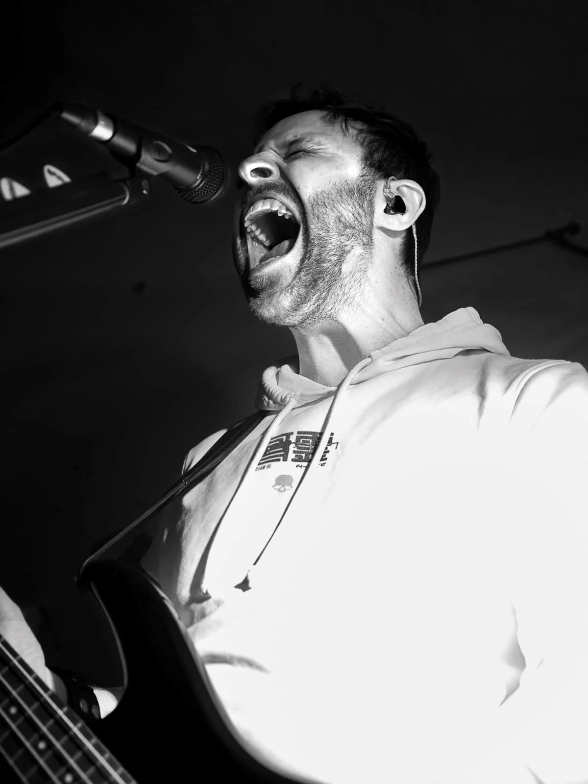 Black and white photo of a man singing into a microphone while playing an electric bass guitar, wearing a hoodie, with an intense expression.