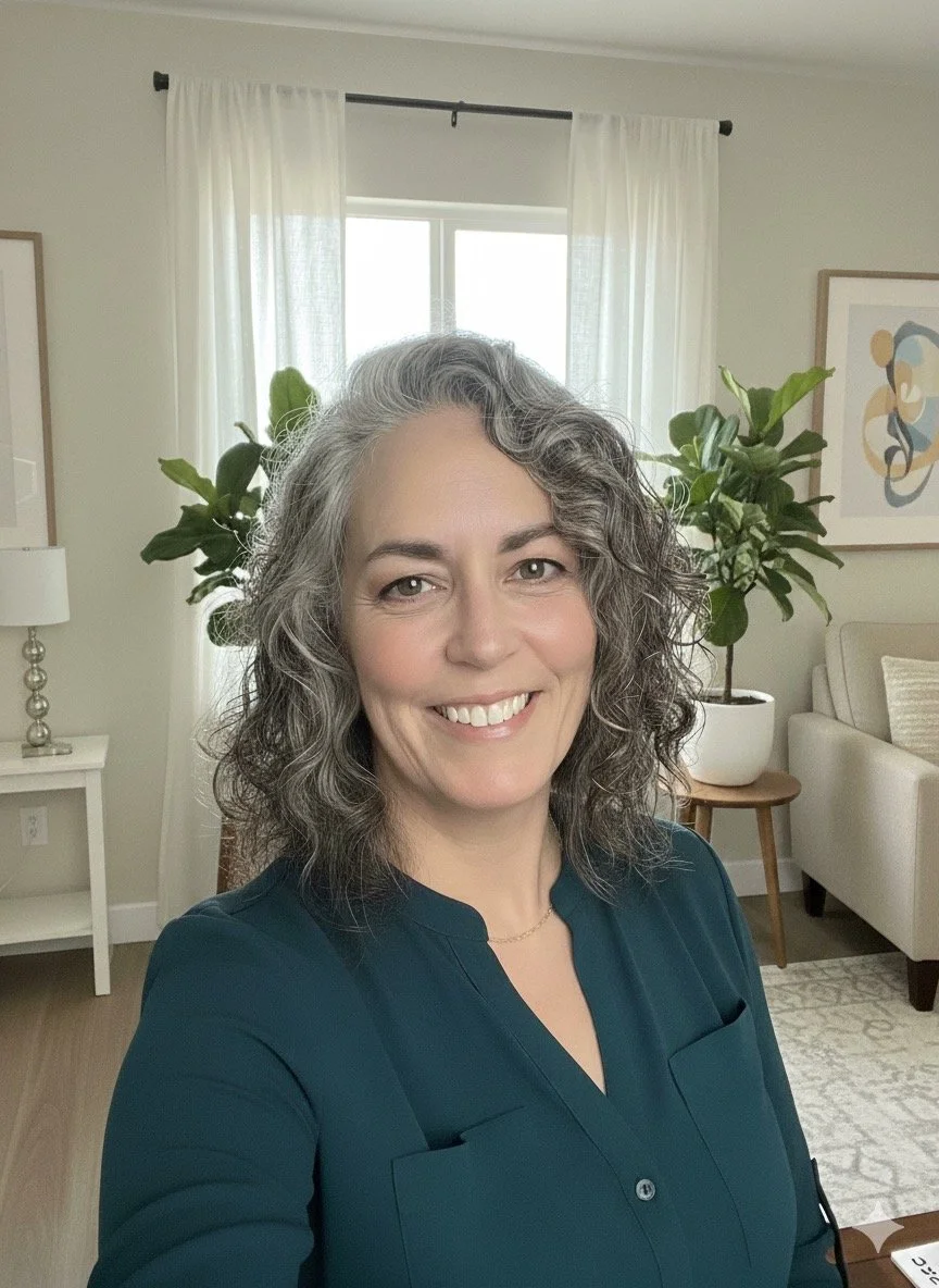 A woman with curly gray hair smiling in a living room with light-colored walls, white curtains, a framed abstract art piece, white sofa, green plants, and a side table with a lamp.