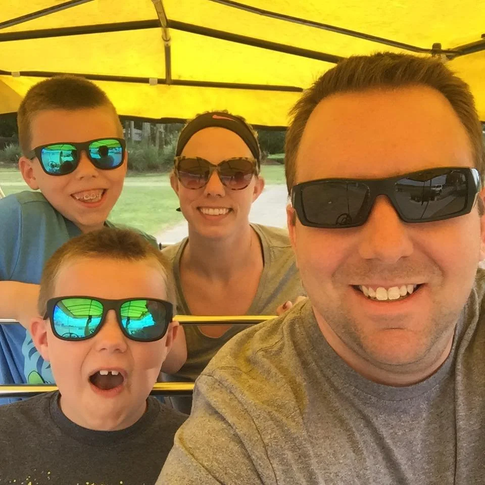 A family of four smiling and wearing sunglasses, taking a selfie under a yellow canopy at an outdoor amusement park.