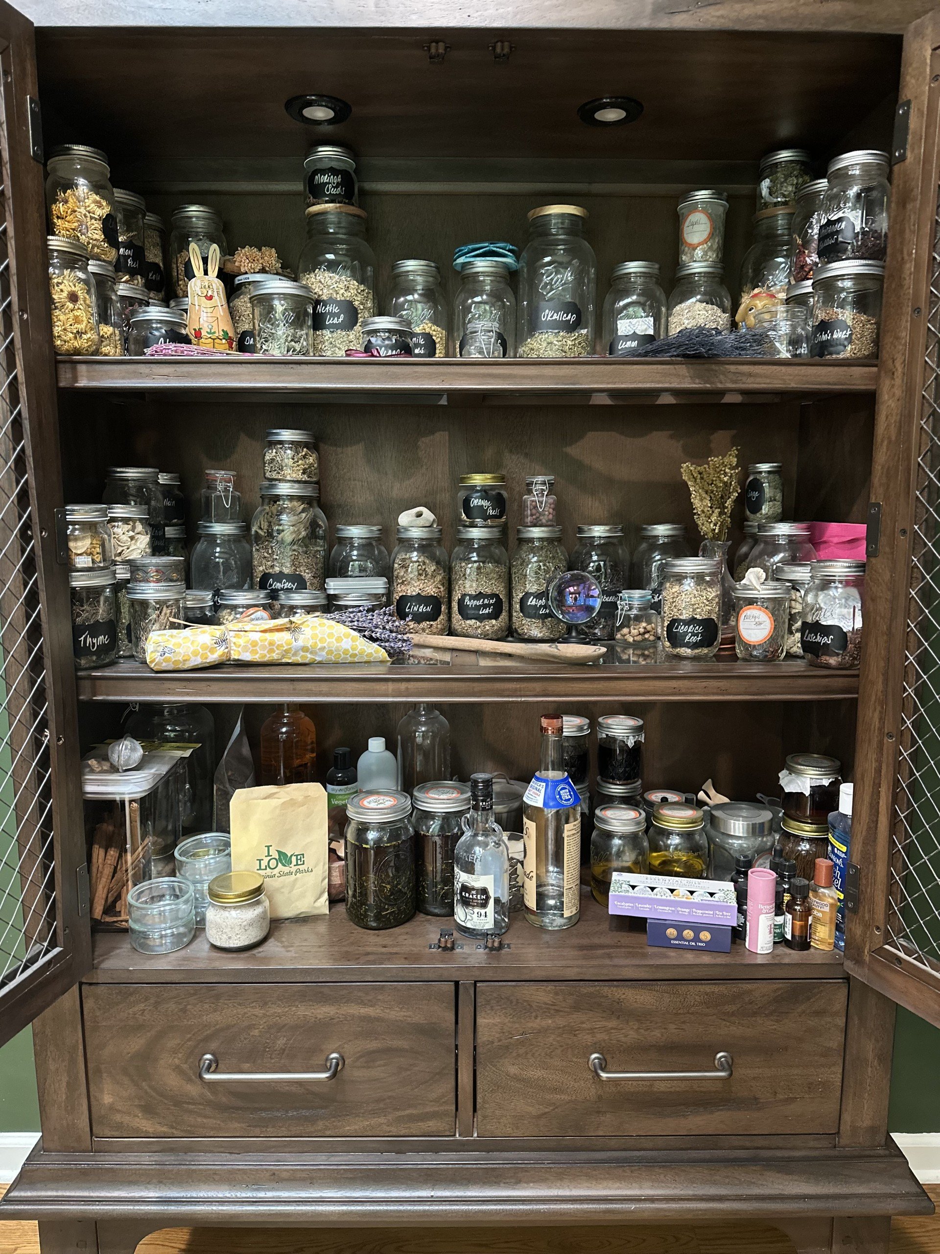 A wooden cabinet filled with glass jars and bottles containing dried herbs, leaves, and spices, with some labeled with handwritten tags, and assorted small bottles and containers on the lower shelves.