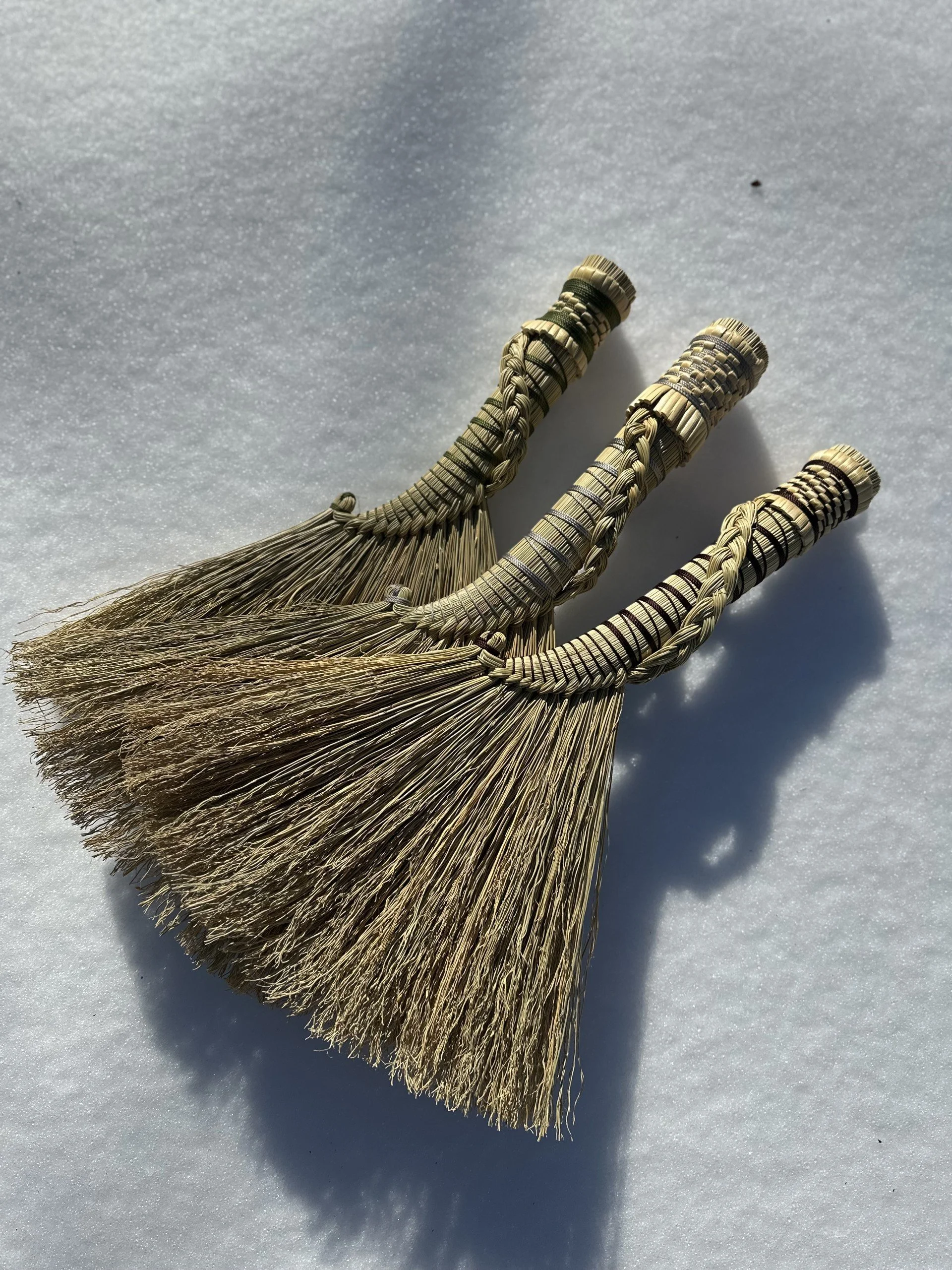 Three traditional broomsticks made of straw with woven handles lying on snow and casting shadows.
