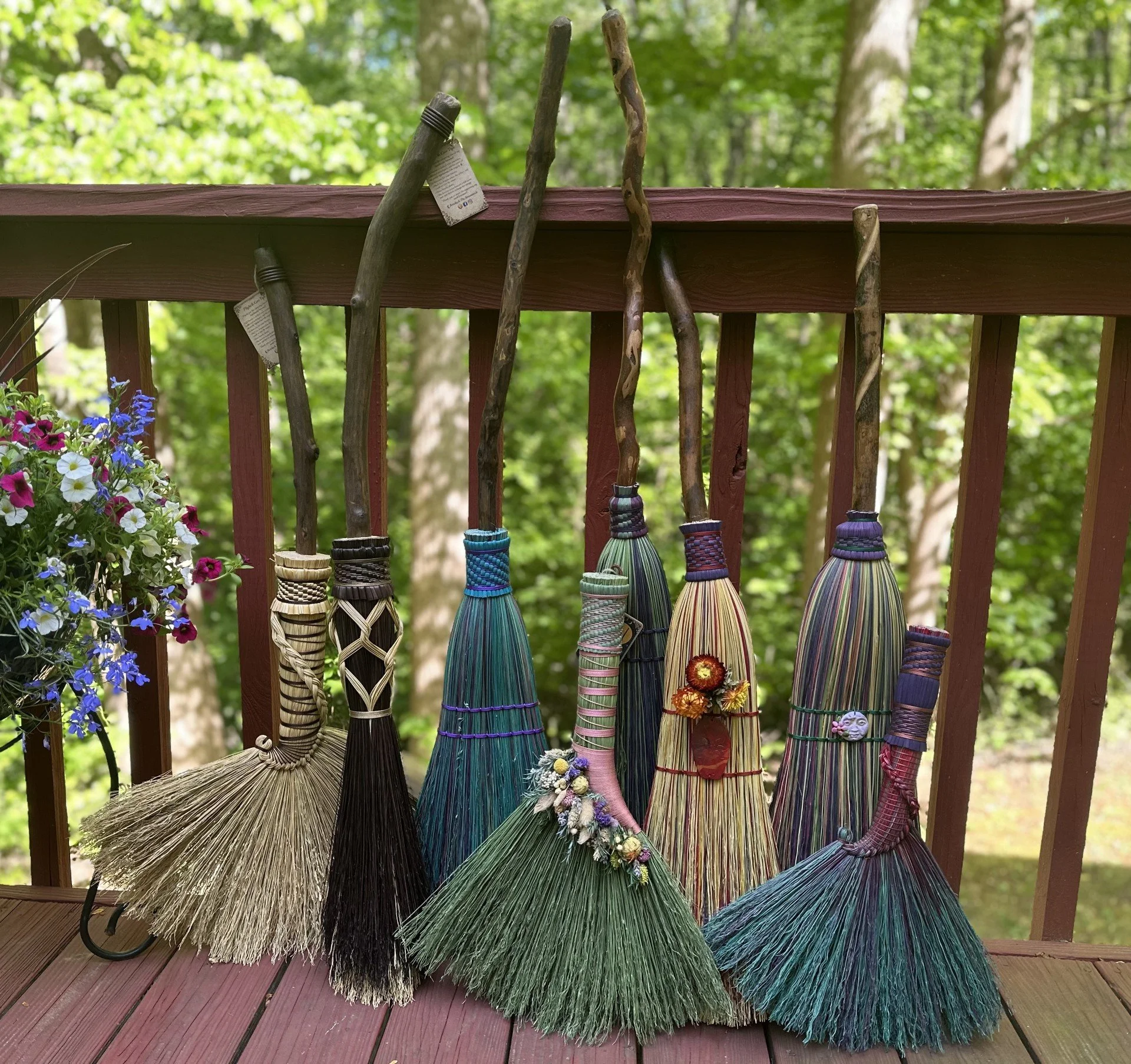 Colorful handmade broomsticks hanging on a wooden railing outdoors with green trees in the background.