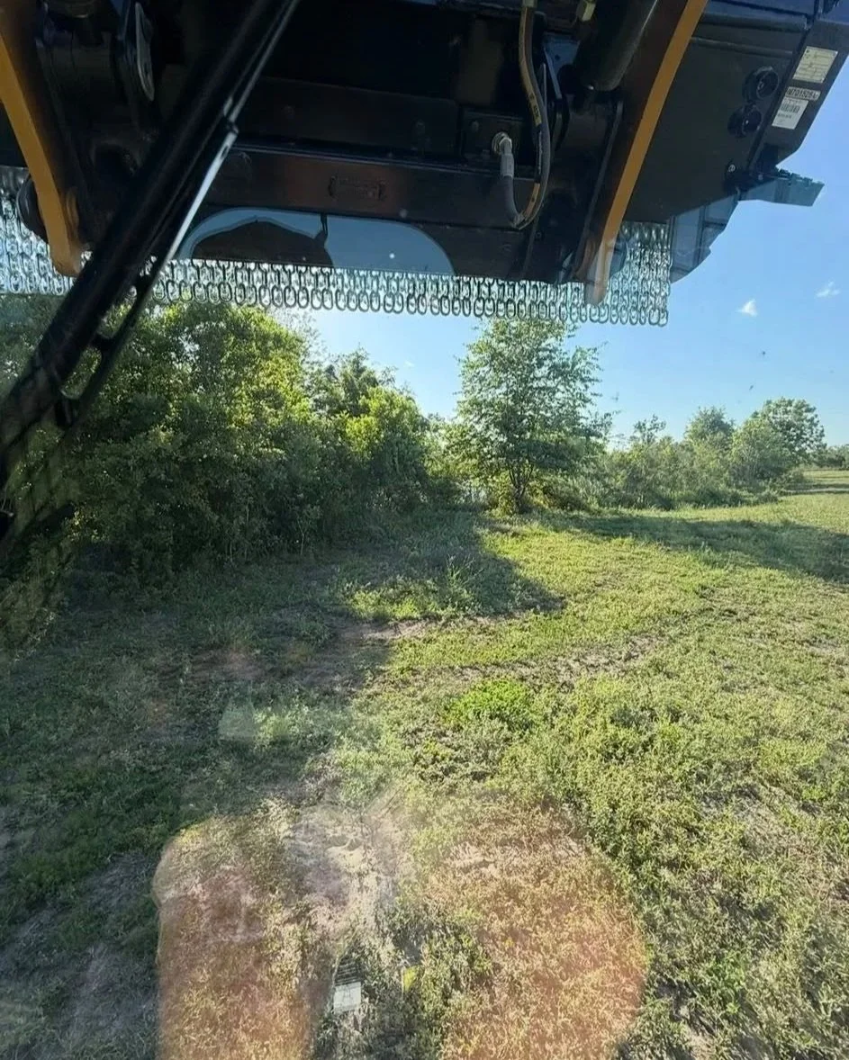 View from inside a piece of heavy construction equipment, looking at green grass, bushes, and trees on a sunny day.