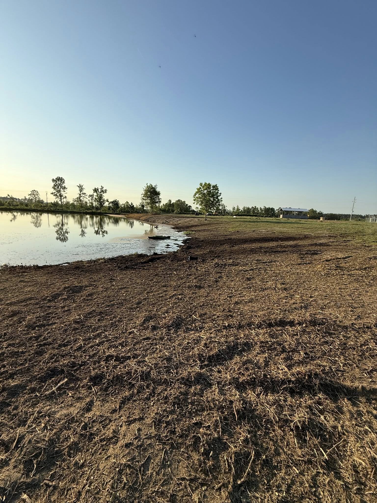 A rural landscape with a body of water, trees along the shoreline, a dirt and grass area, a small building in the distance, and a clear blue sky with some airplanes flying overhead.