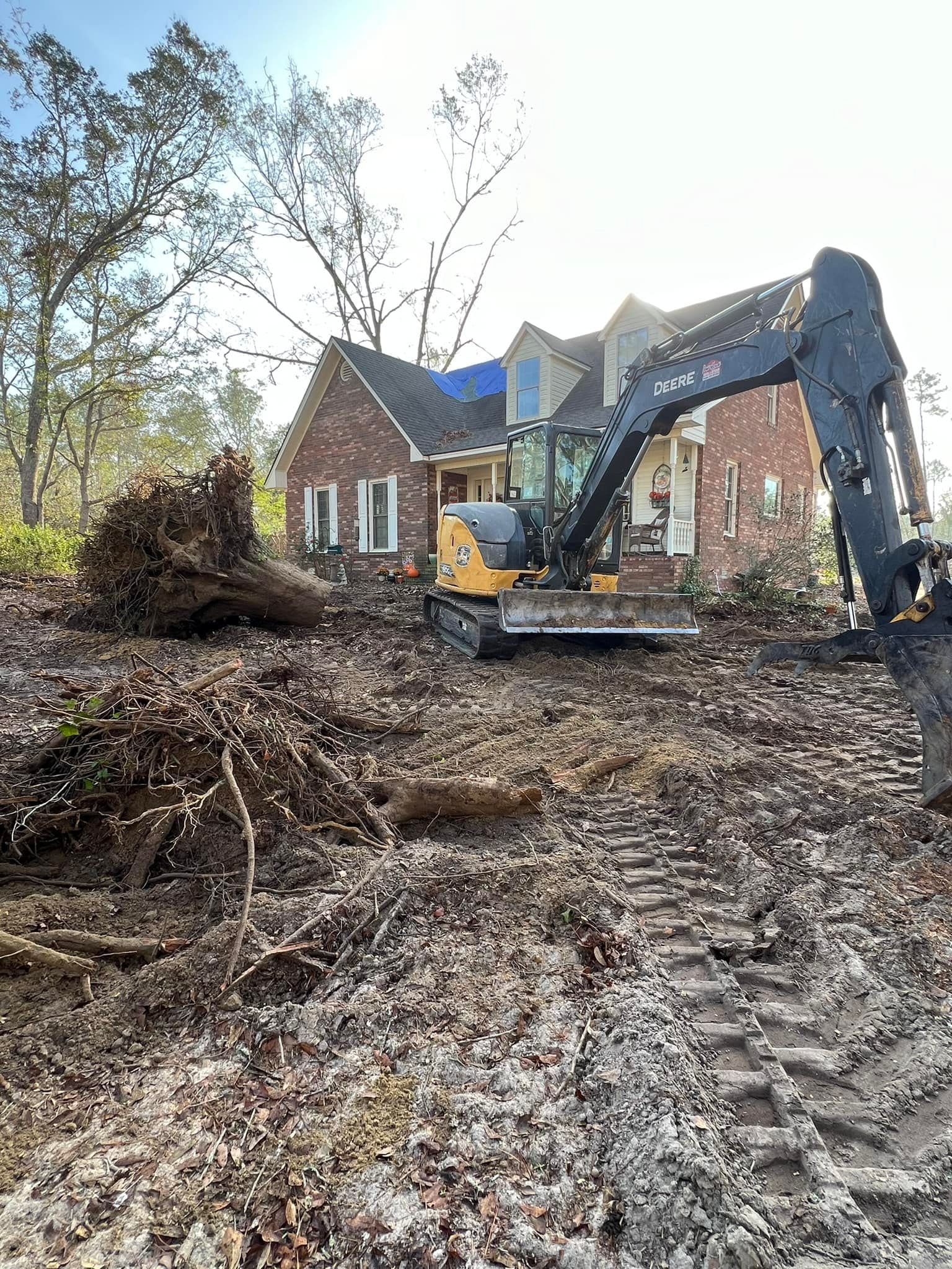 A construction site in front of a brick house with trees around. A small yellow and black excavator is moving soil and tree roots. There are visible tracks in the dirt from the excavator and a fallen tree with roots on the ground.