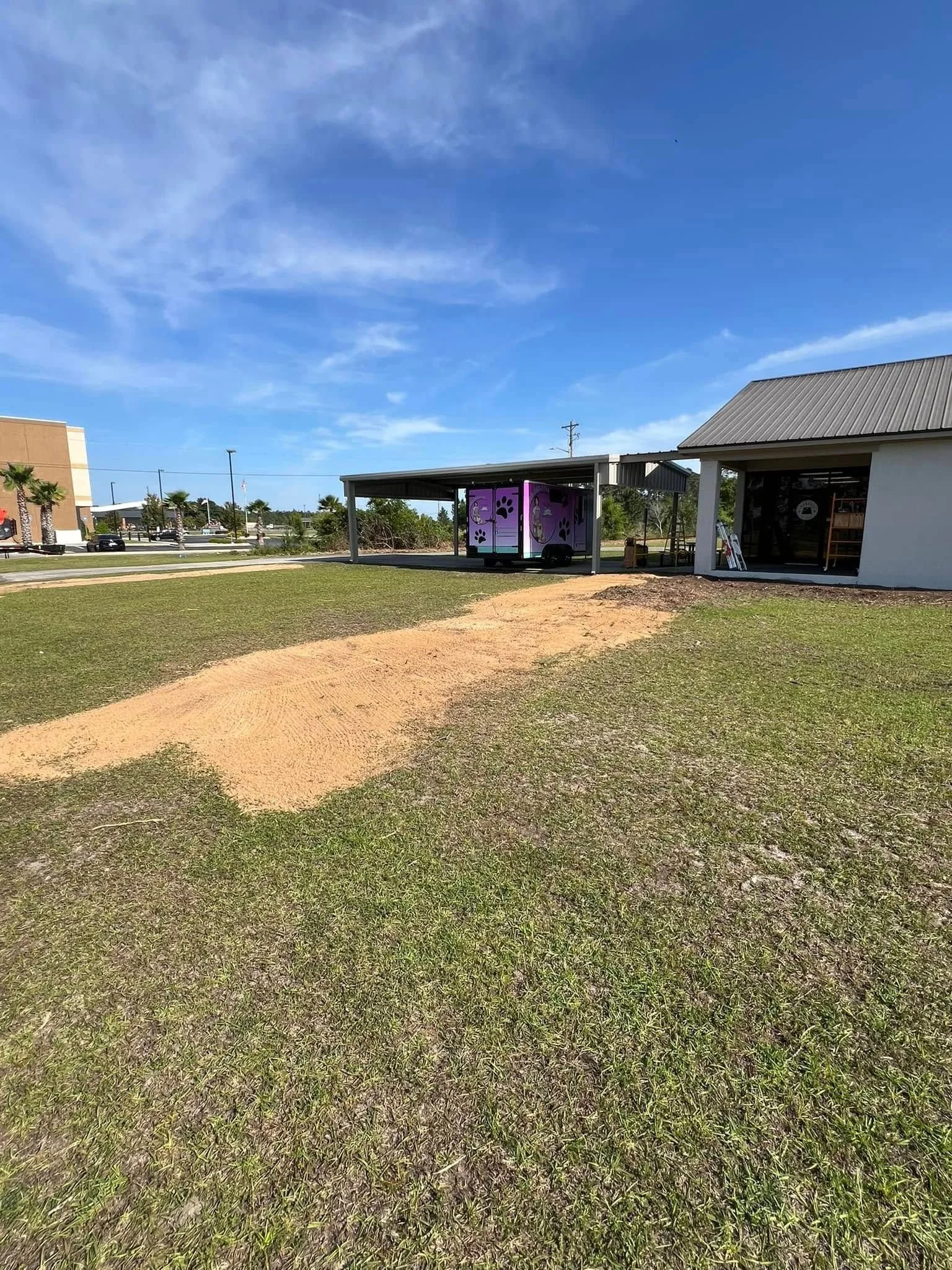 An outdoor area with green grass, a dirt path, a small covered structure with a purple cart decorated with paw prints, and a building with a metal roof under a partly cloudy sky.