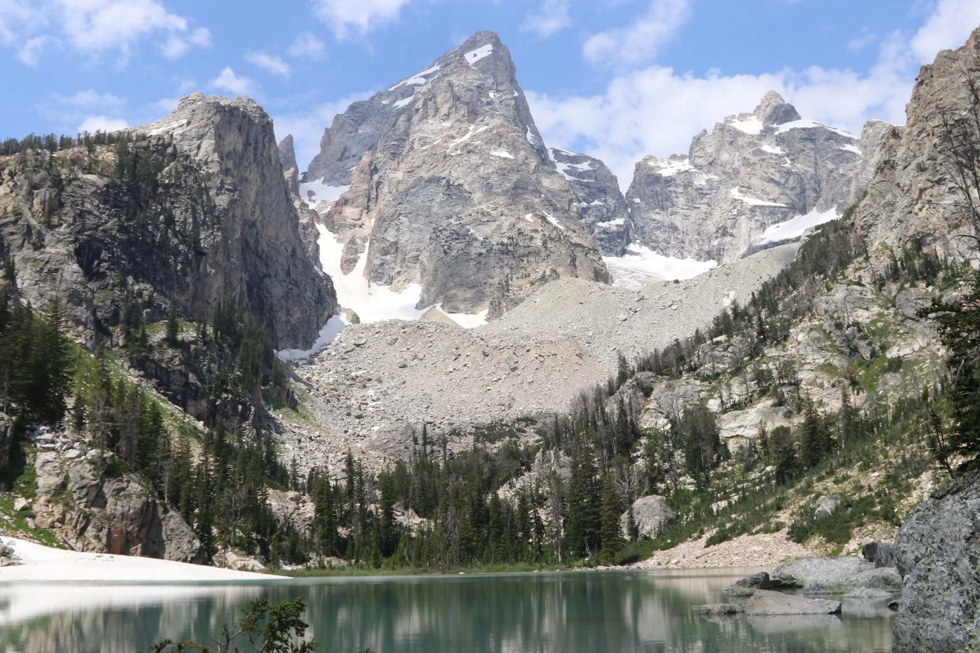 A mountain landscape featuring tall, rugged peaks with patches of snow, dense pine trees at the base, and a calm lake reflecting the scenery under a partly cloudy sky.