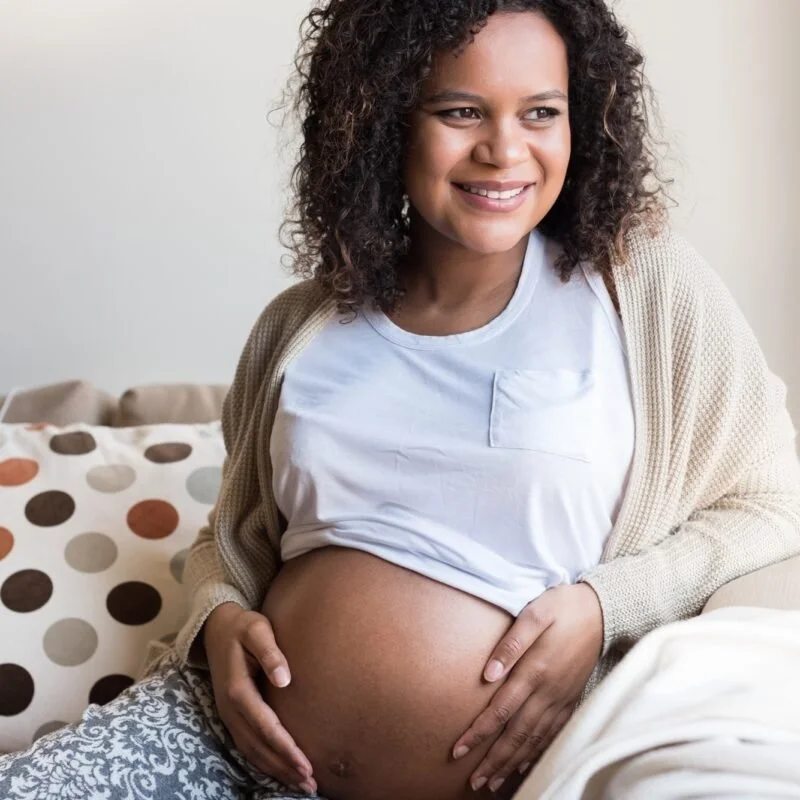 Smiling pregnant woman sitting on a couch with her hand on her belly, in a cozy room.