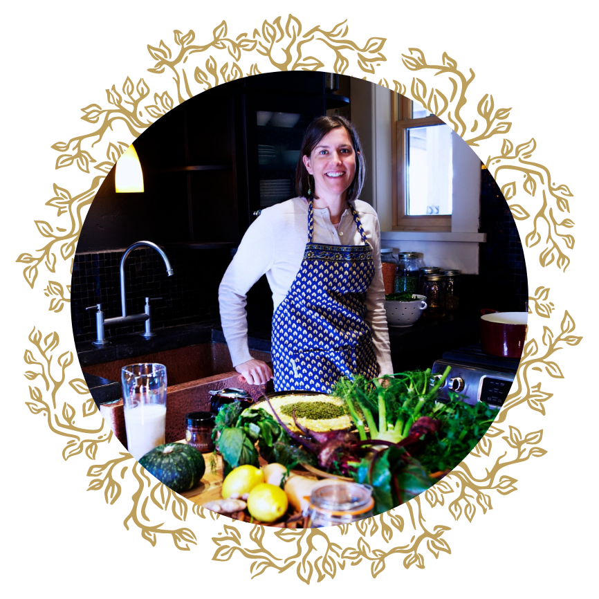 A woman in a kitchen wearing a blue apron, smiling, surrounded by fresh vegetables and cooking ingredients on the counter.
