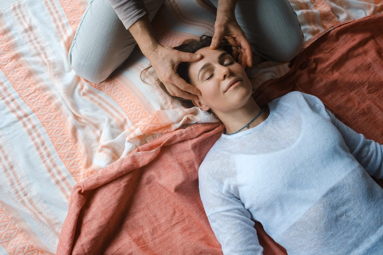 A woman lying on a bed with her eyes closed, receiving a head massage from a person kneeling beside her. The bed has patterned sheets and blankets.