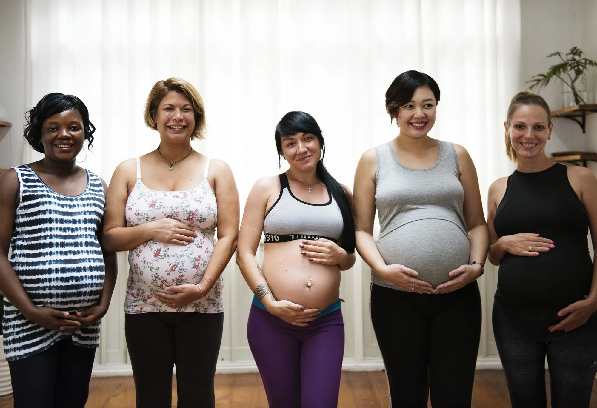 A diverse group of six women, some pregnant, standing together indoors in front of white curtains with a wooden floor and small shelves with a plant in the background, smiling.
