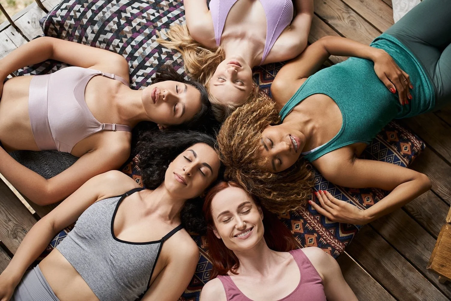Six women lying on a wooden floor in a circle with their heads together and eyes closed, relaxed and smiling.