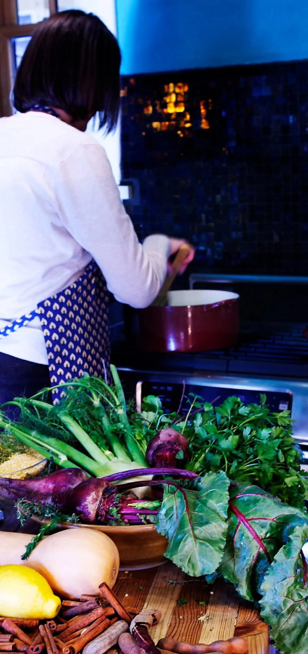 Person cooking in a kitchen with fresh vegetables, including beets, leafy greens, a lemon, butternut squash, and cinnamon sticks on a wooden table.