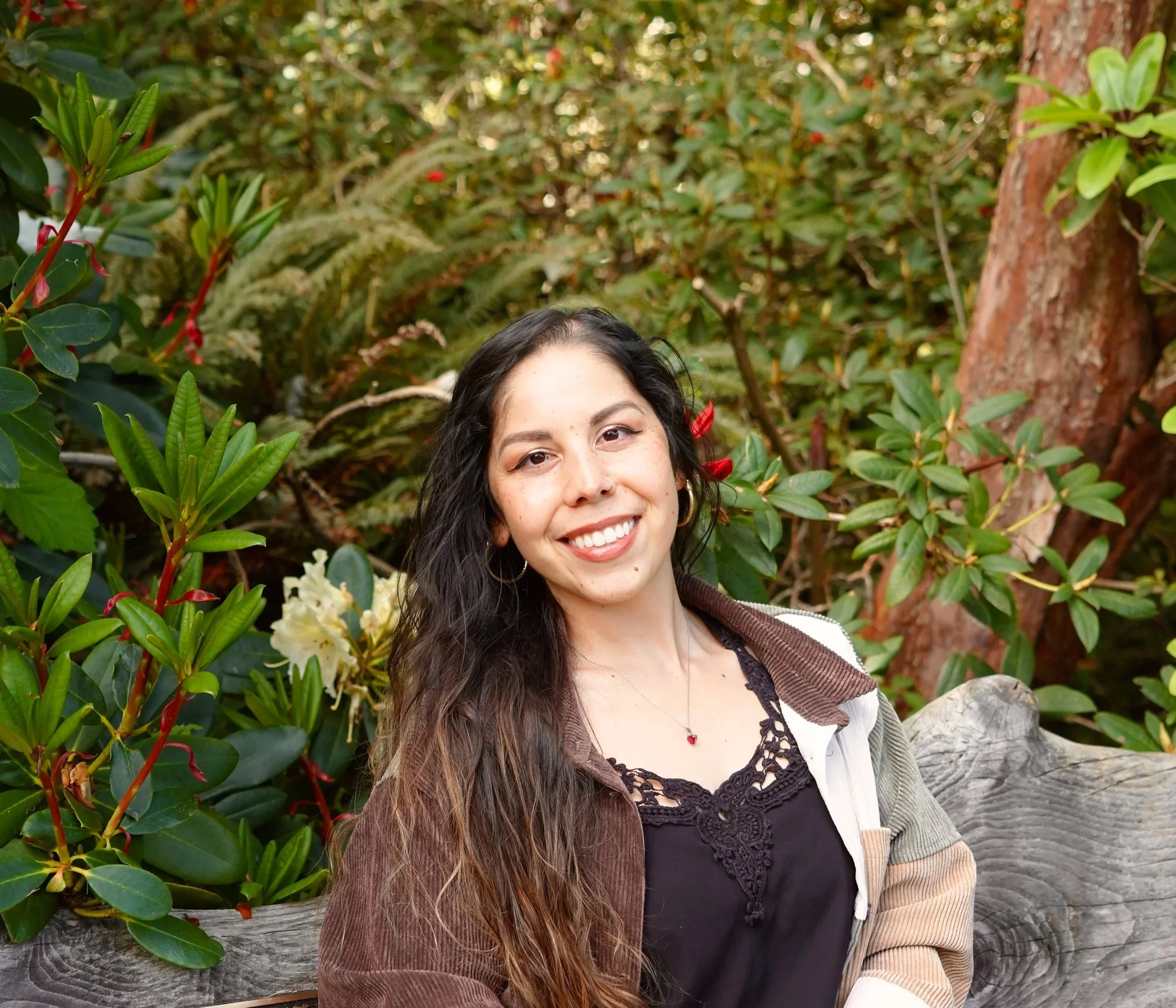 A close-up of a smiling woman with long curly hair, wearing a black lace top, a gray cardigan, and a red heart necklace, standing outdoors among green plants and a tree.