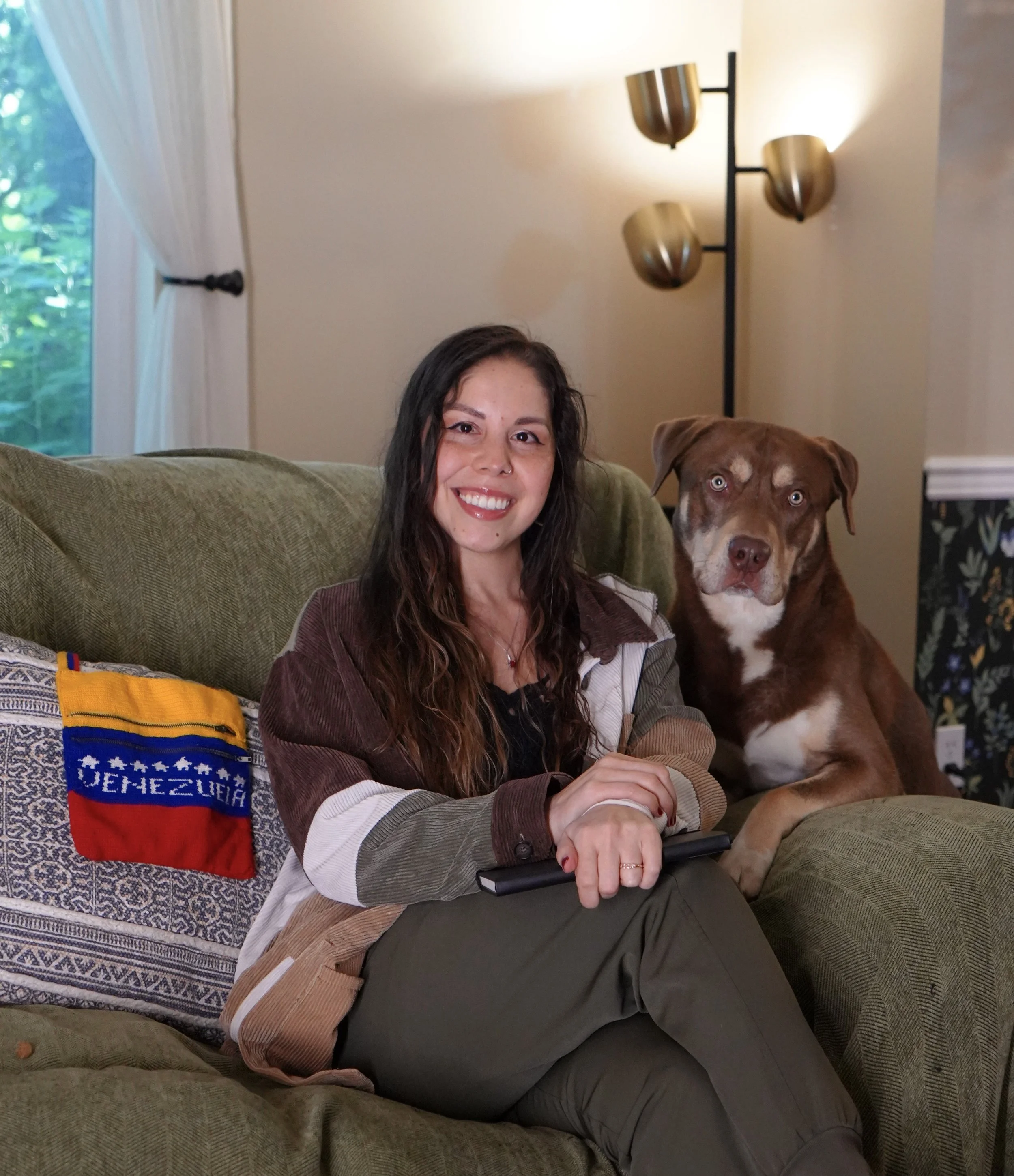 A woman with long dark curly hair and light skin is sitting on a green upholstered couch, smiling at the camera. Beside her, a large brown dog with white markings on its chest and bright blue eyes is lying down. In the background, there is a window with white curtains and a black metal floor lamp with three gold-colored shades. An item with the flag of Venezuela is on the left side of the couch.