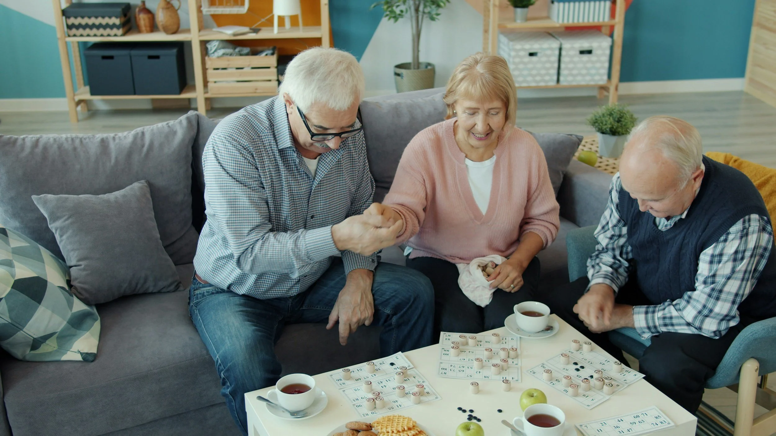 Three elderly people playing bingo at a living room table with drinks and snacks.