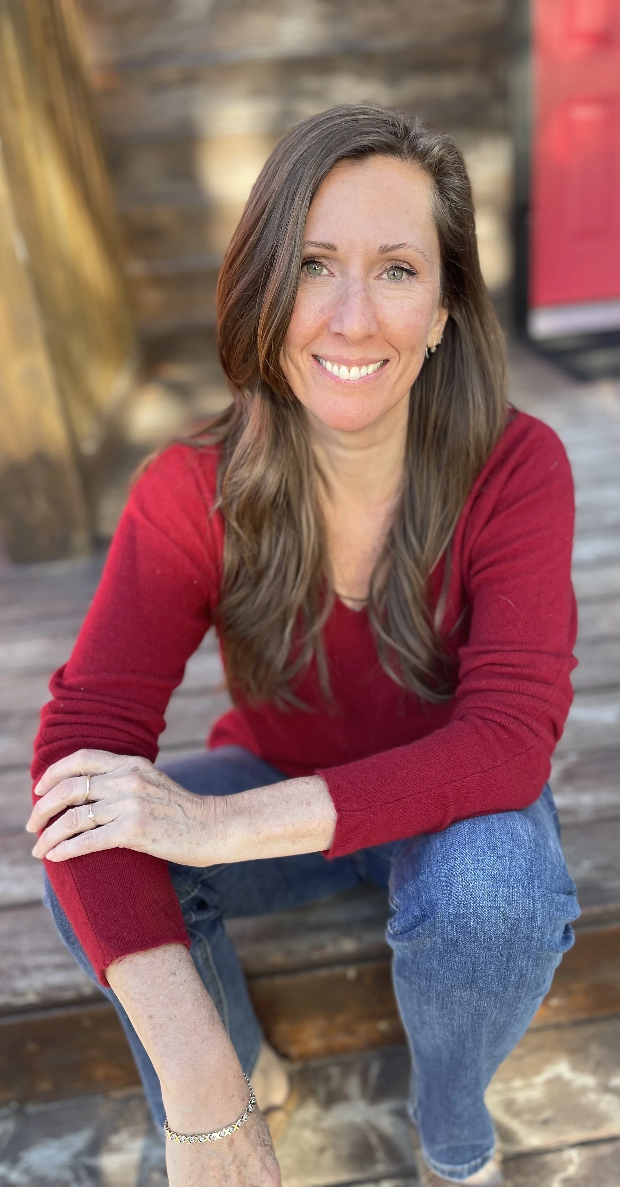 A woman with long brown hair and green eyes wearing a red long-sleeve shirt, sitting on wooden stairs outdoors, smiling at the camera.