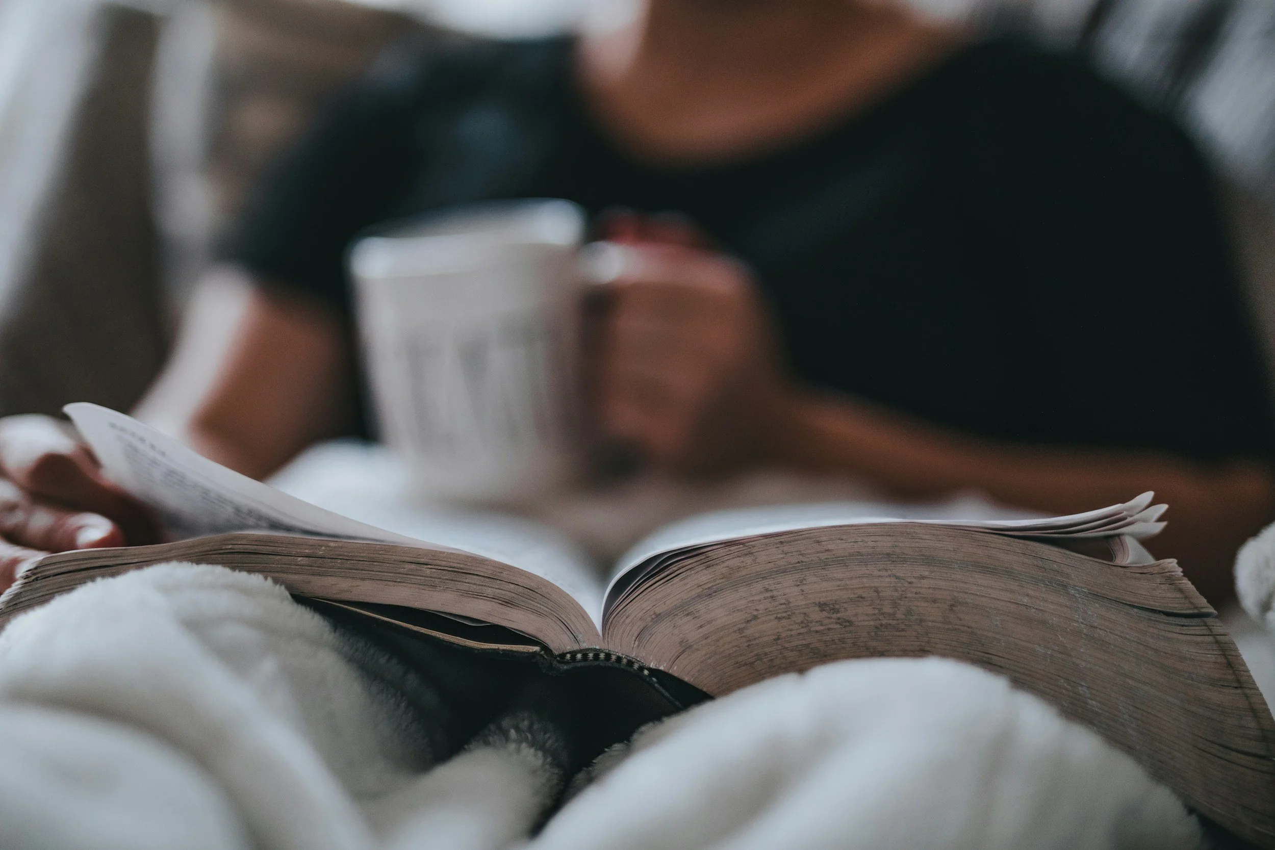 A person is sitting on a soft surface, reading a book with a slightly open spine. The focus is on the open pages of the book, with the person's hand visible at the side. The background is blurred, and the person is wearing dark clothing.
