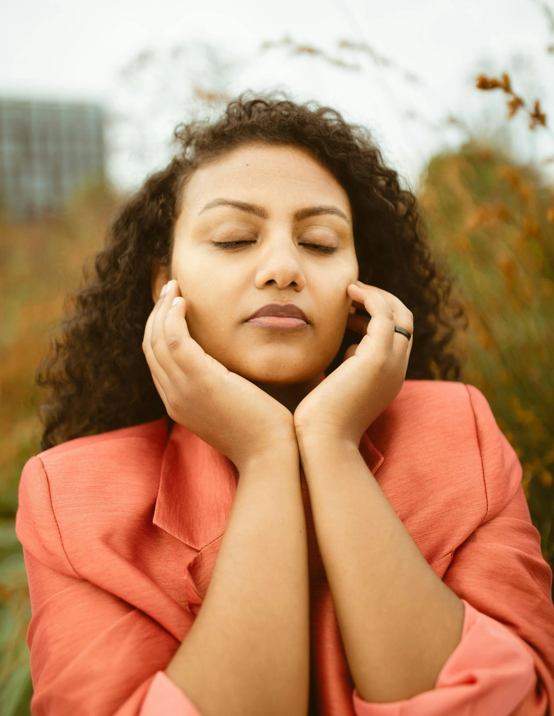A woman with curly hair and closed eyes, wearing a pink blazer, with her hands gently touching her face outdoors.